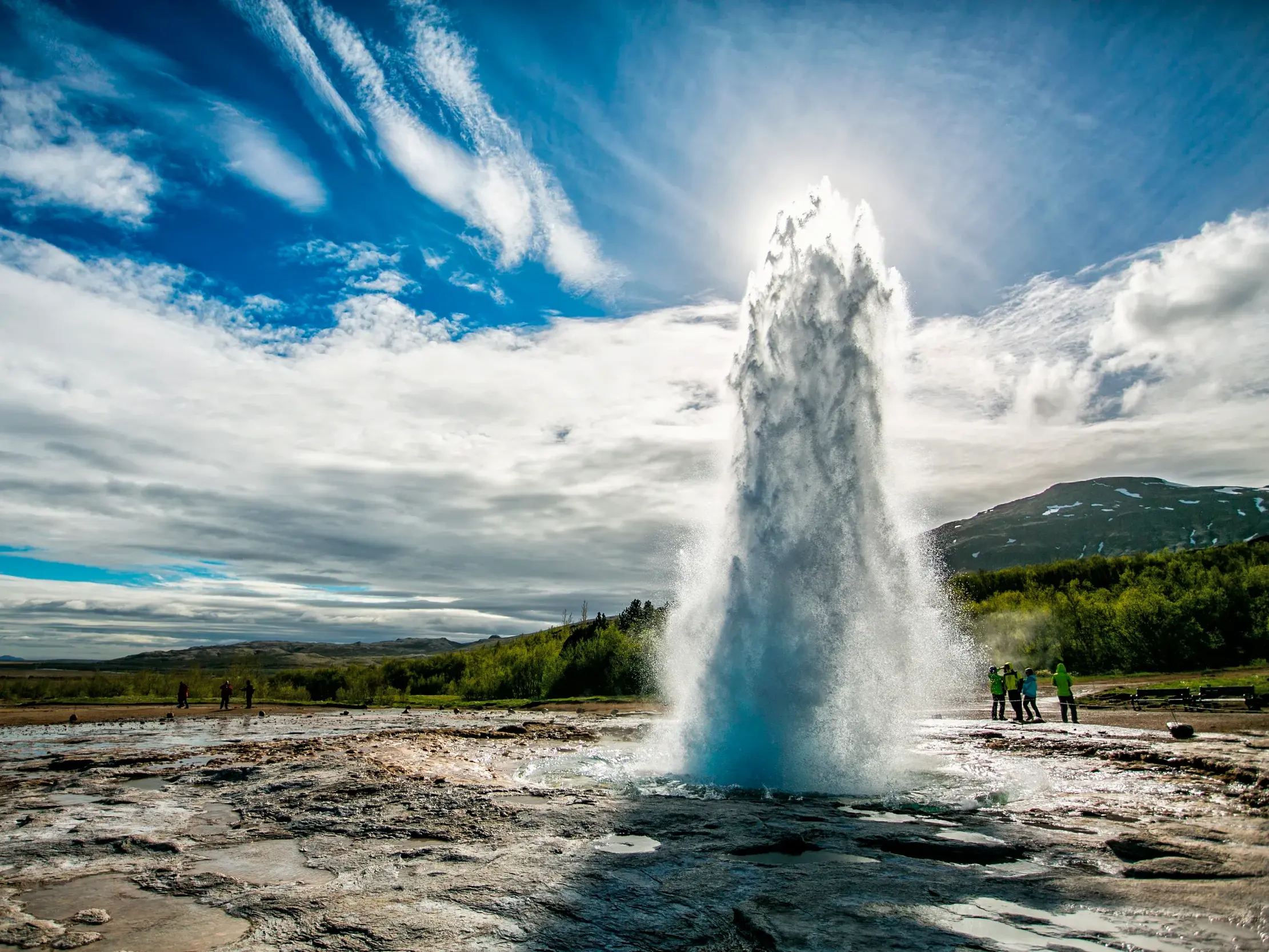 Eine kleine Reisegruppe beobachtet die gewaltige Wasserfontäne des ausbrechenden Geysirs Strokkur auf dem Golden Circle in Island.