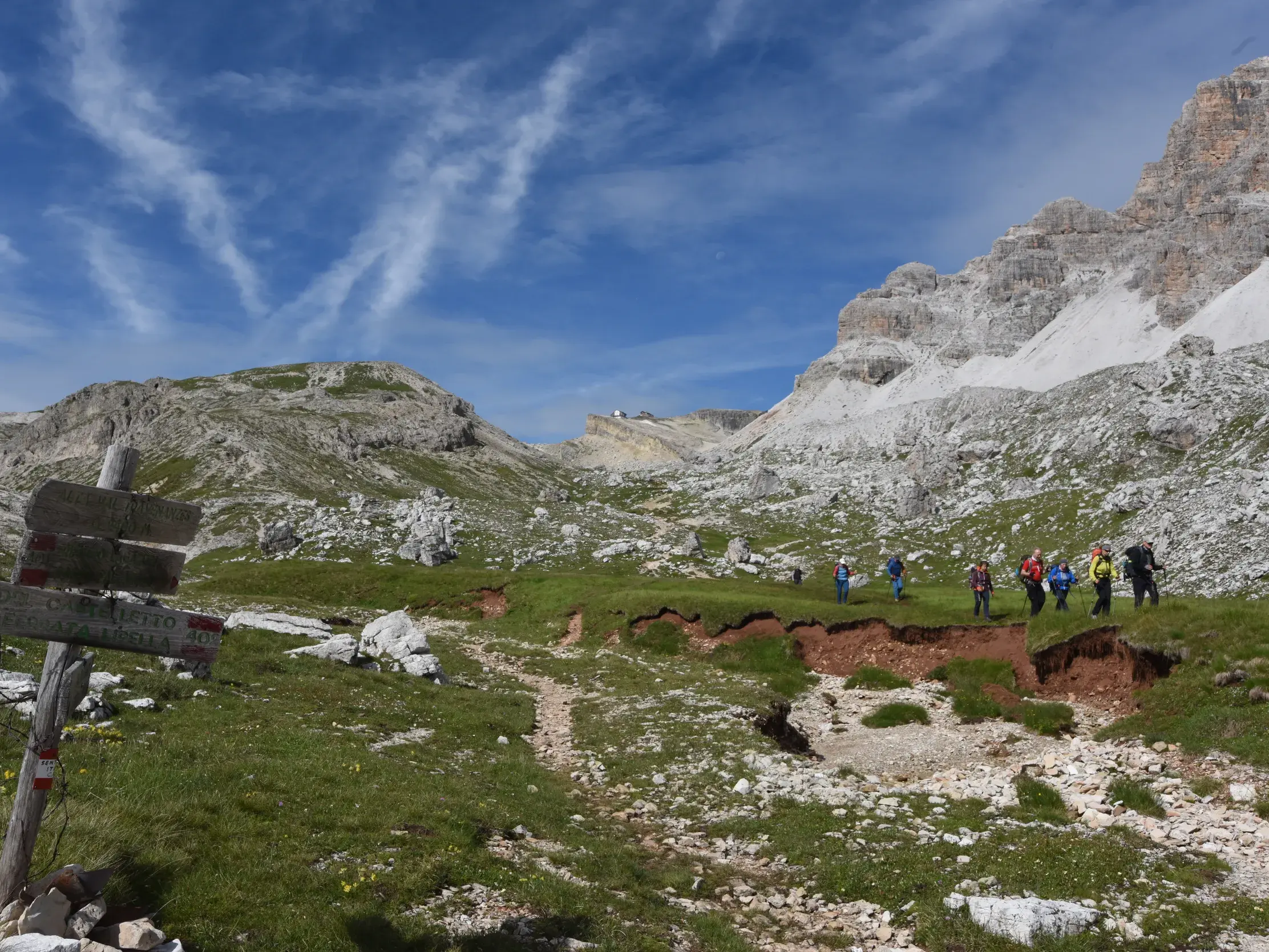 Hölzerner Wegweiser auf einem Wanderweg in den Dolomiten, im Hintergrund eine Hauser Wandergruppe auf einer Alm vor imposanten Gipfeln.