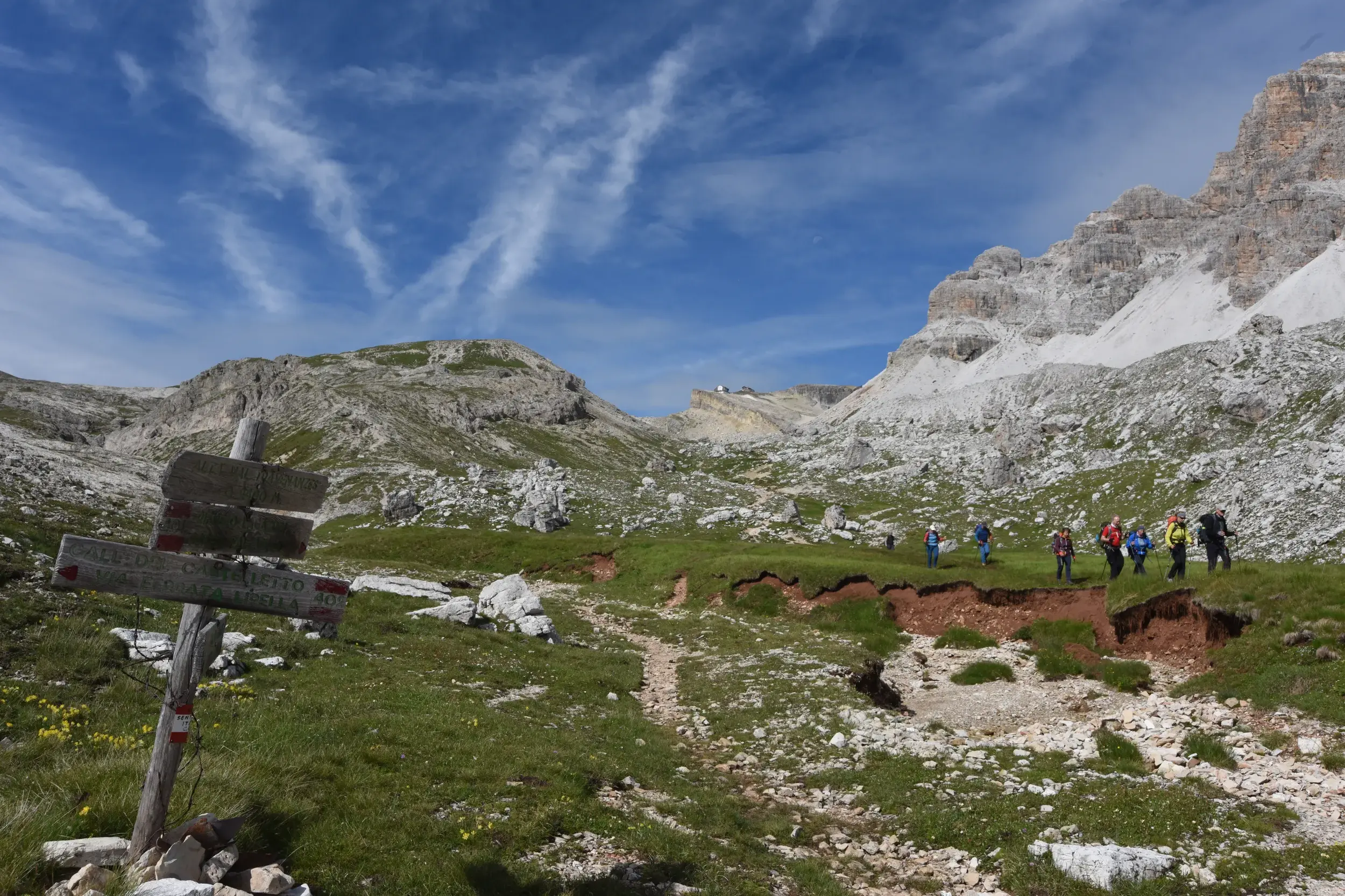Hölzerner Wegweiser auf einem Wanderweg in den Dolomiten, im Hintergrund eine Hauser Wandergruppe auf einer Alm vor imposanten Gipfeln.