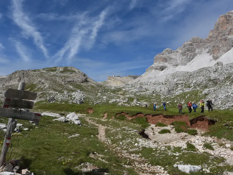Hölzerner Wegweiser auf einem Wanderweg in den Dolomiten, im Hintergrund eine Hauser Wandergruppe auf einer Alm vor imposanten Gipfeln.