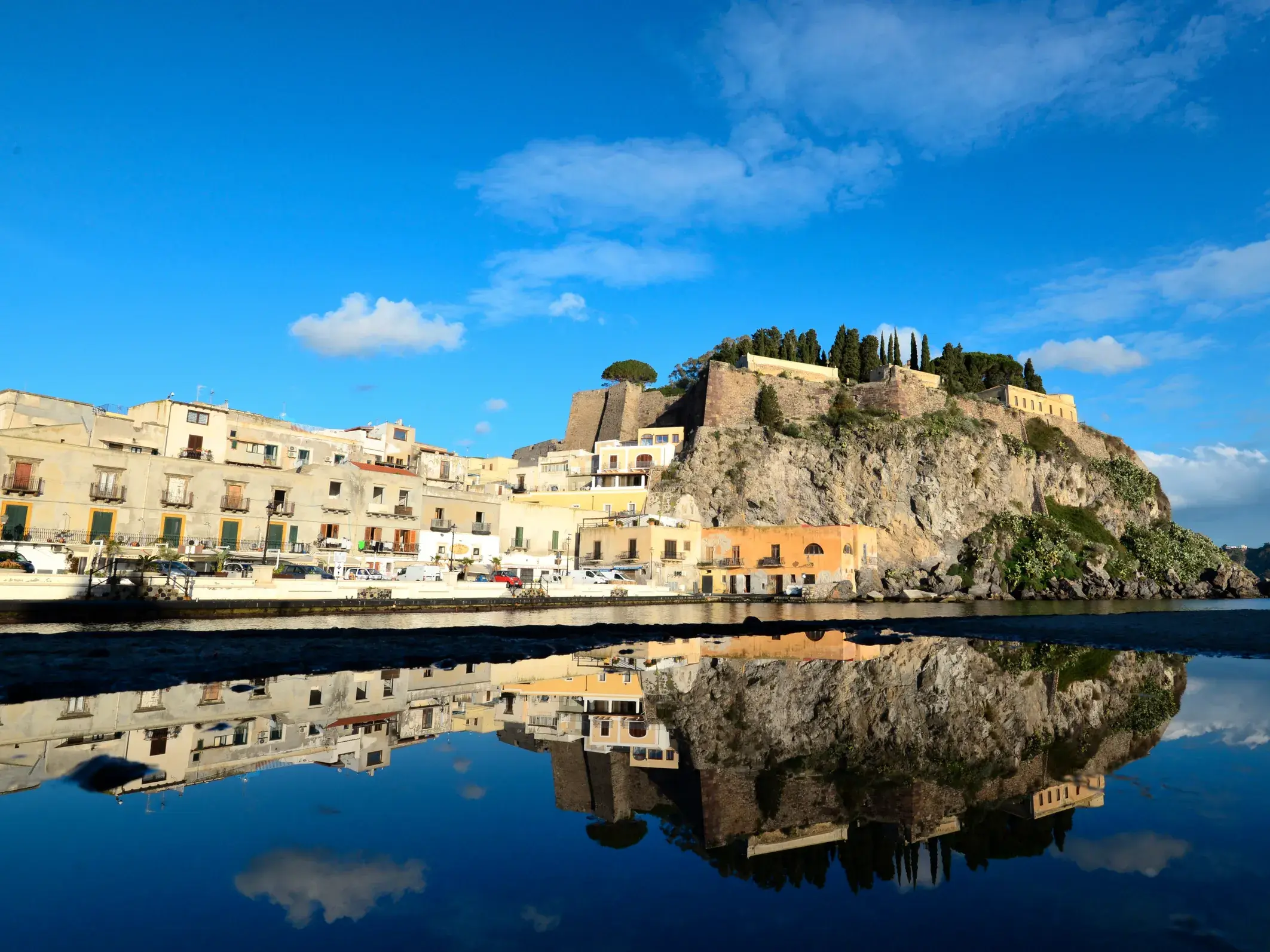 Die befestigte Altstadt von Lipari auf einem Felsen am Meer, die sich bei blauem Himmel perfekt im ruhigen Wasser spiegelt.