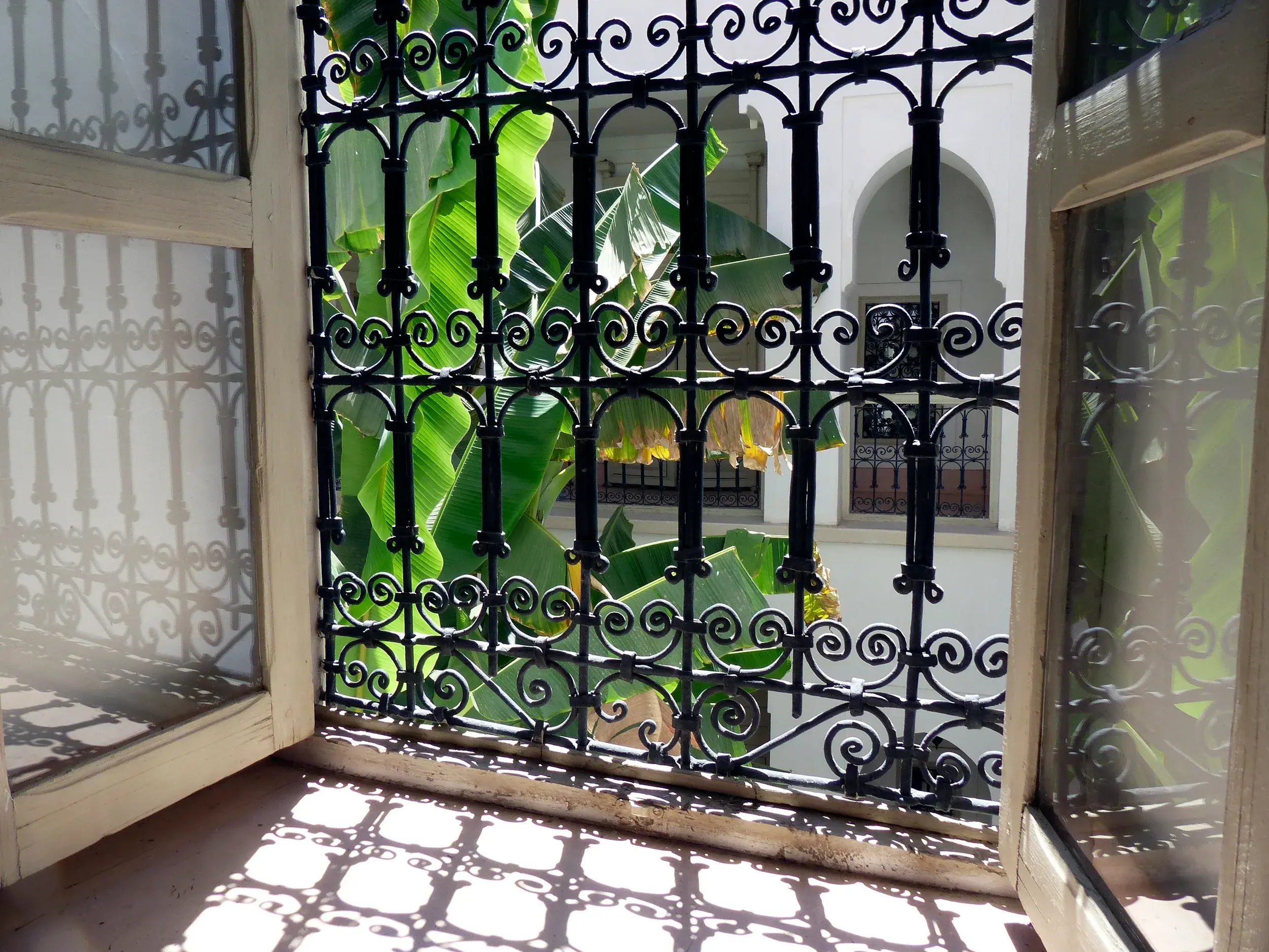 Sonniger Blick durch ein schmiedeeisernes Fenstergitter in den begrünten Innenhof eines traditionellen Riads in Marokko.