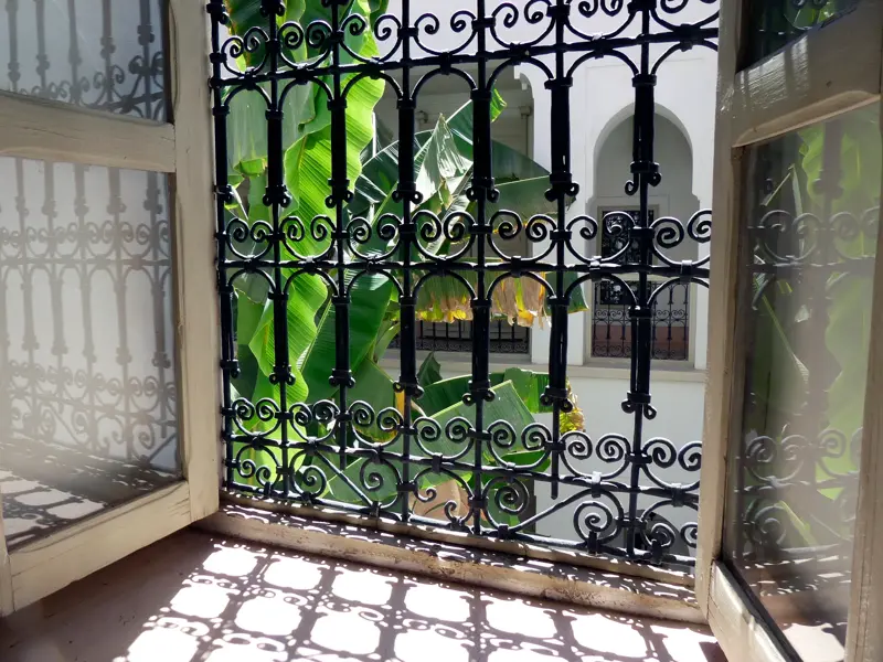 Sonniger Blick durch ein schmiedeeisernes Fenstergitter in den begrünten Innenhof eines traditionellen Riads in Marokko.