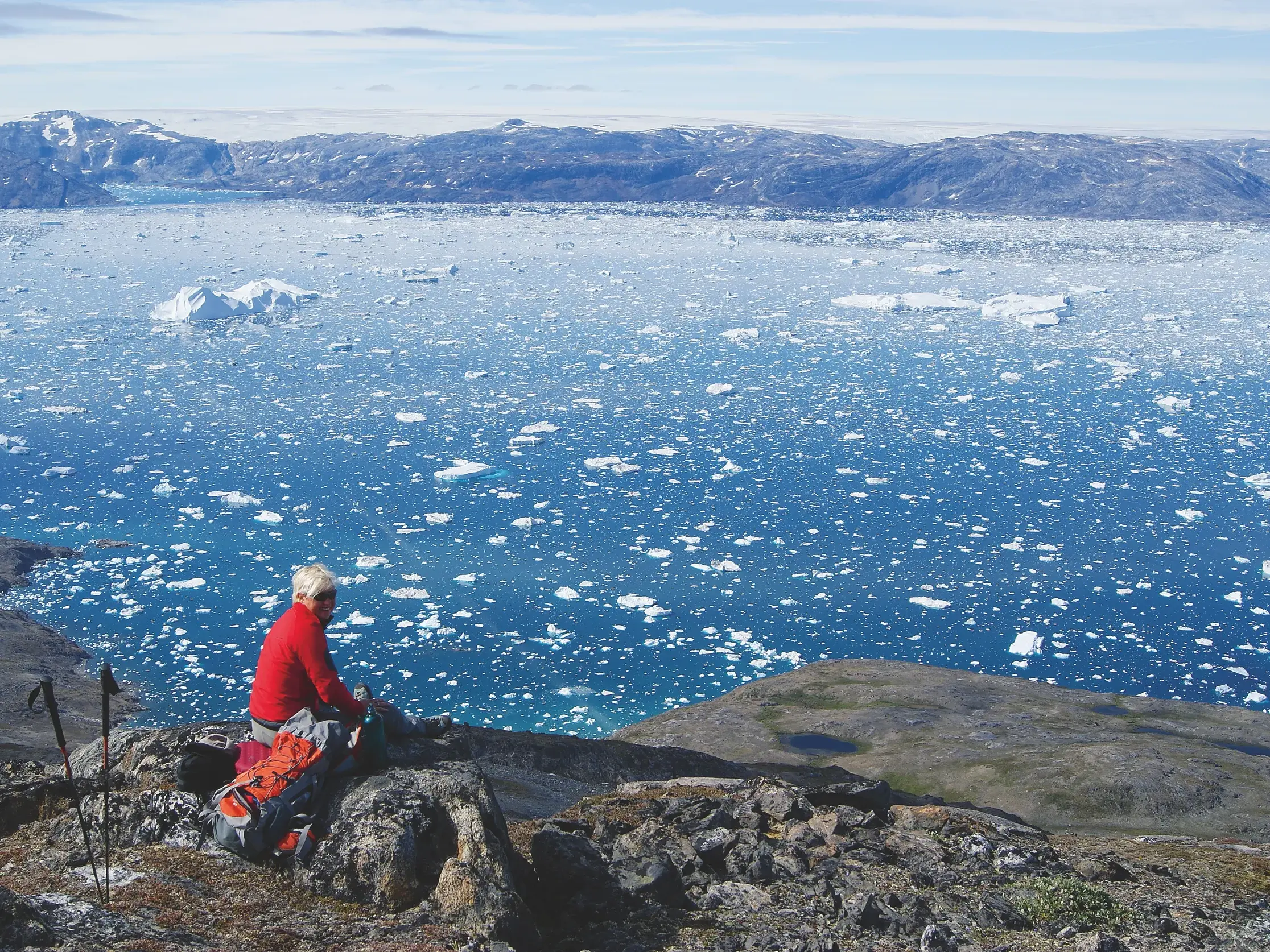 Wanderin ruht bei einer Trekking-Tour auf einem Felsen und blickt über den weiten, eisgefüllten Sermilik-Fjord in Ostgrönland.