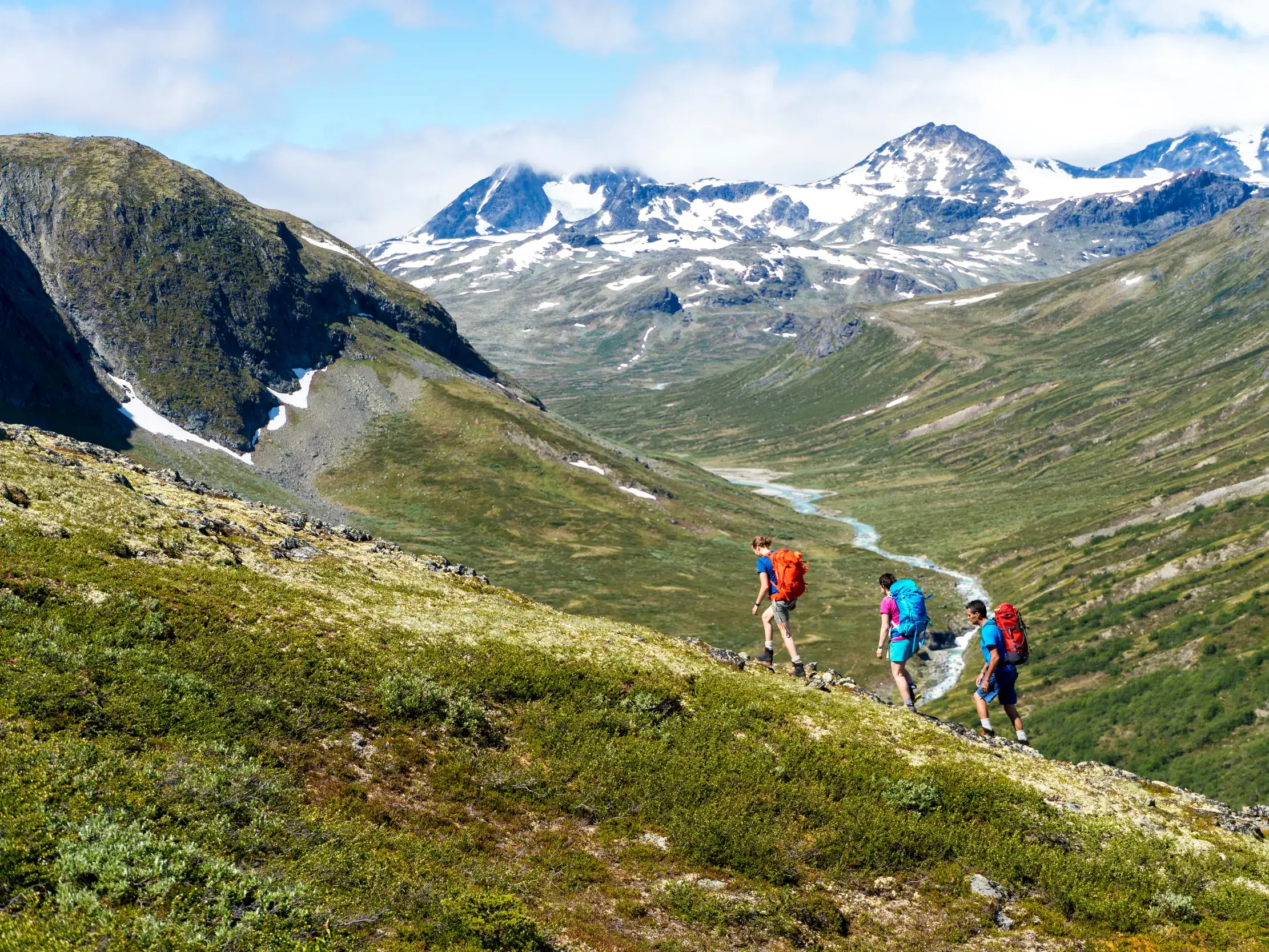 Eine kleine Wandergruppe beim Trekking durch ein weites Bergtal im Jotunheimen Nationalpark mit Blick auf schneebedeckte Gipfel.