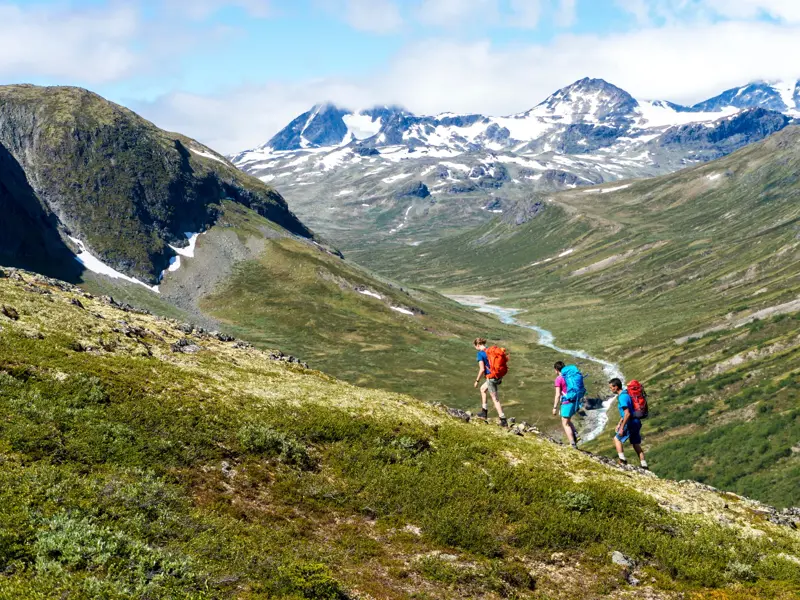 Eine kleine Wandergruppe beim Trekking durch ein weites Bergtal im Jotunheimen Nationalpark mit Blick auf schneebedeckte Gipfel.