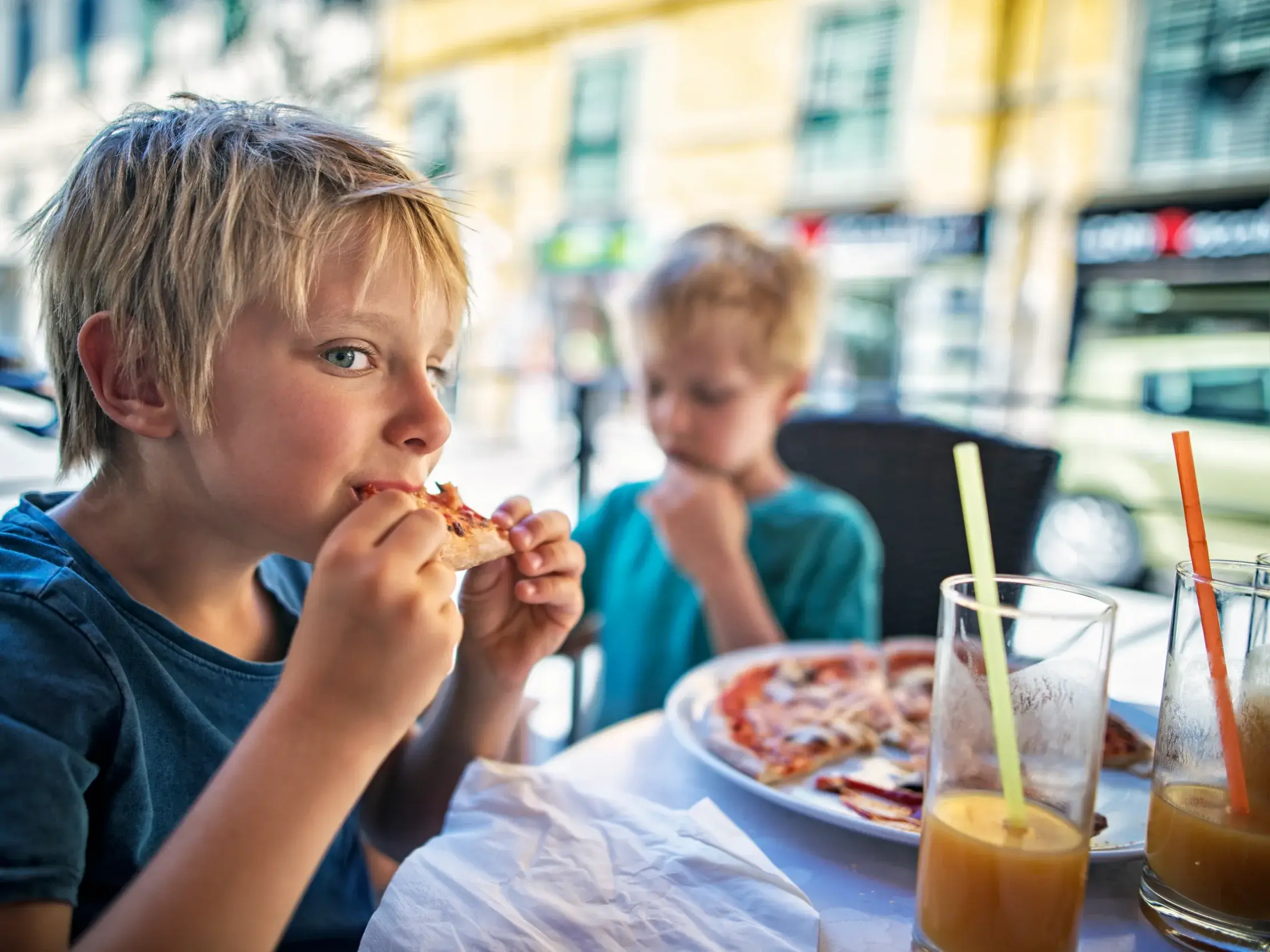 Blonder Junge blickt in die Kamera, während er in einer Trattoria in Italien genussvoll ein Stück Pizza isst.
