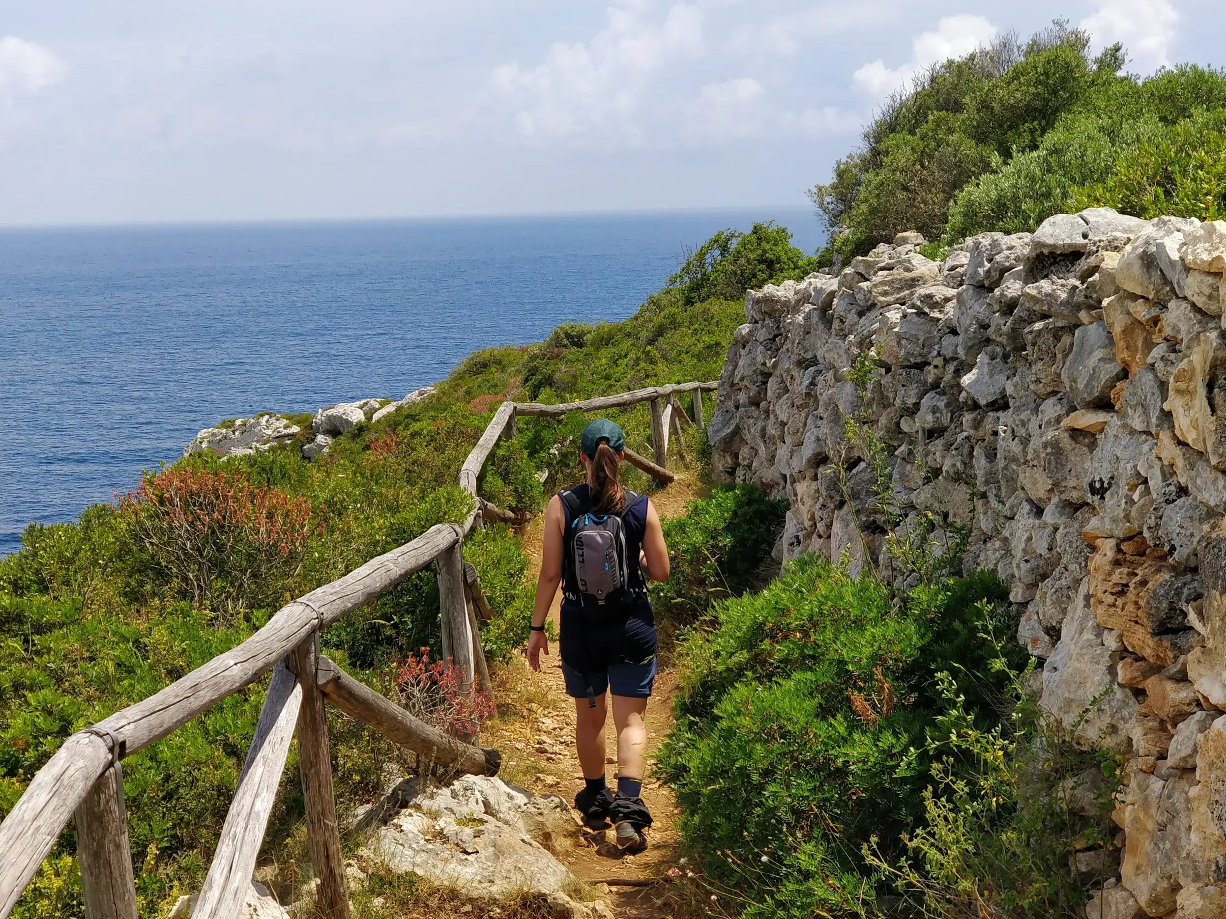 Eine Frau wandert auf einem schmalen Küstenpfad in Apulien, gesäumt von einer Steinmauer und mit Blick auf das blaue Meer.