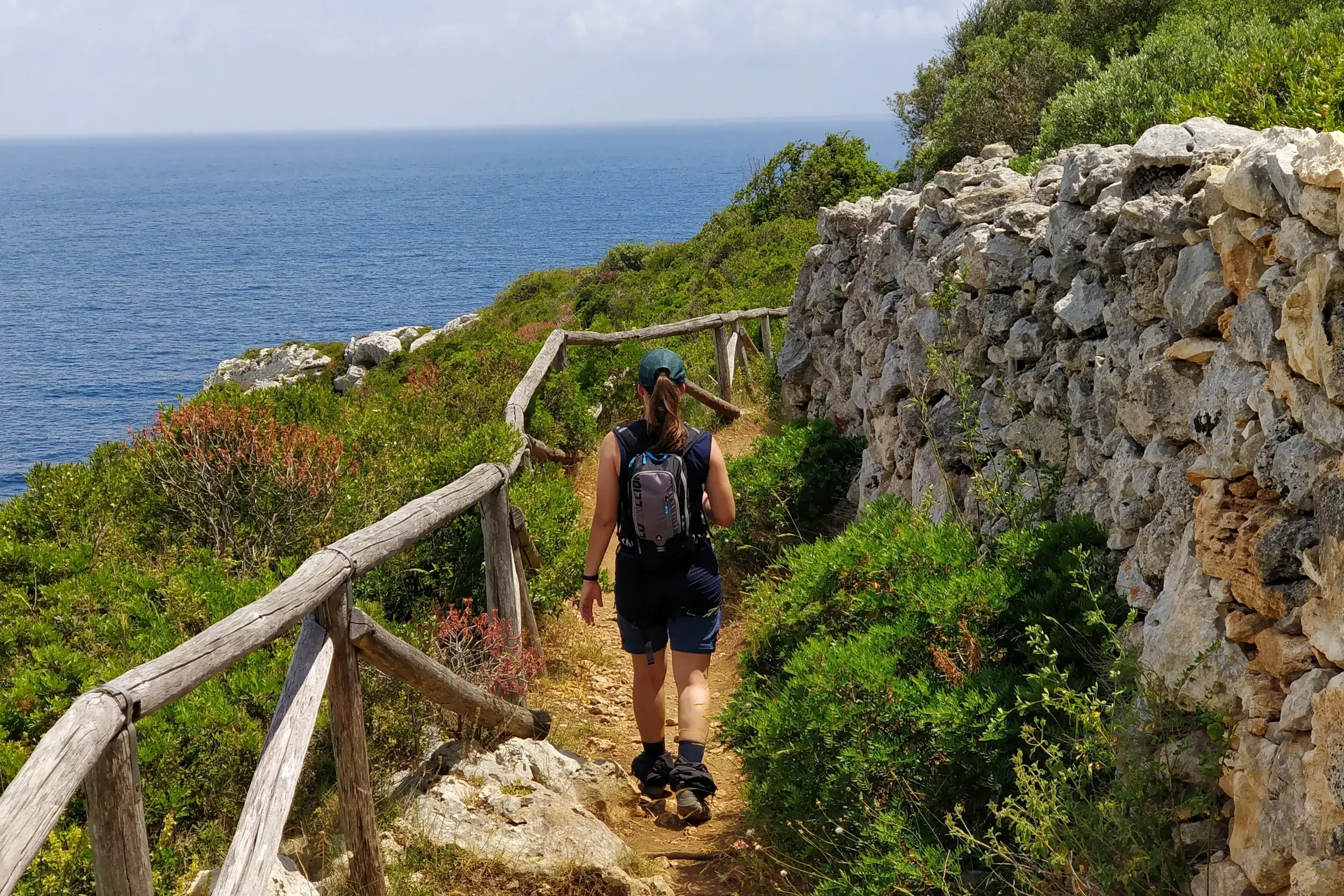 Eine Frau wandert auf einem schmalen Küstenpfad in Apulien, gesäumt von einer Steinmauer und mit Blick auf das blaue Meer.