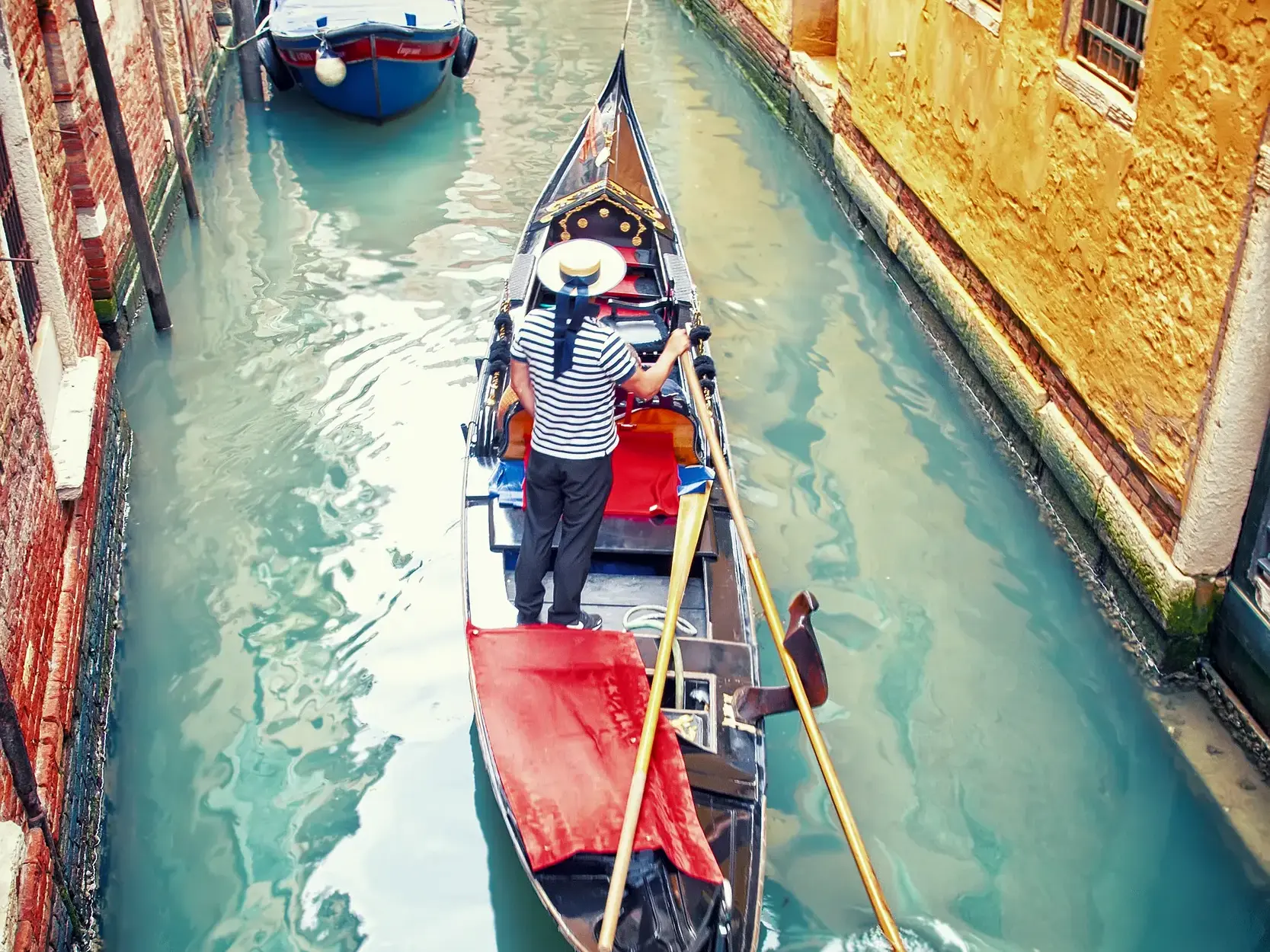 Ein Gondoliere in traditioneller Kleidung steuert seine Gondel von oben gesehen durch einen engen Kanal zwischen alten Häusern in Venedig.