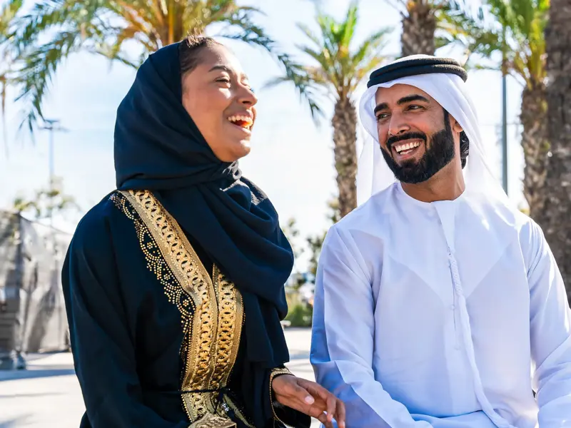 Frau in Abaya und Mann in Thawb lachen bei einer herzlichen Begegnung auf einer Studienreise in Saudi-Arabien unter Palmen.