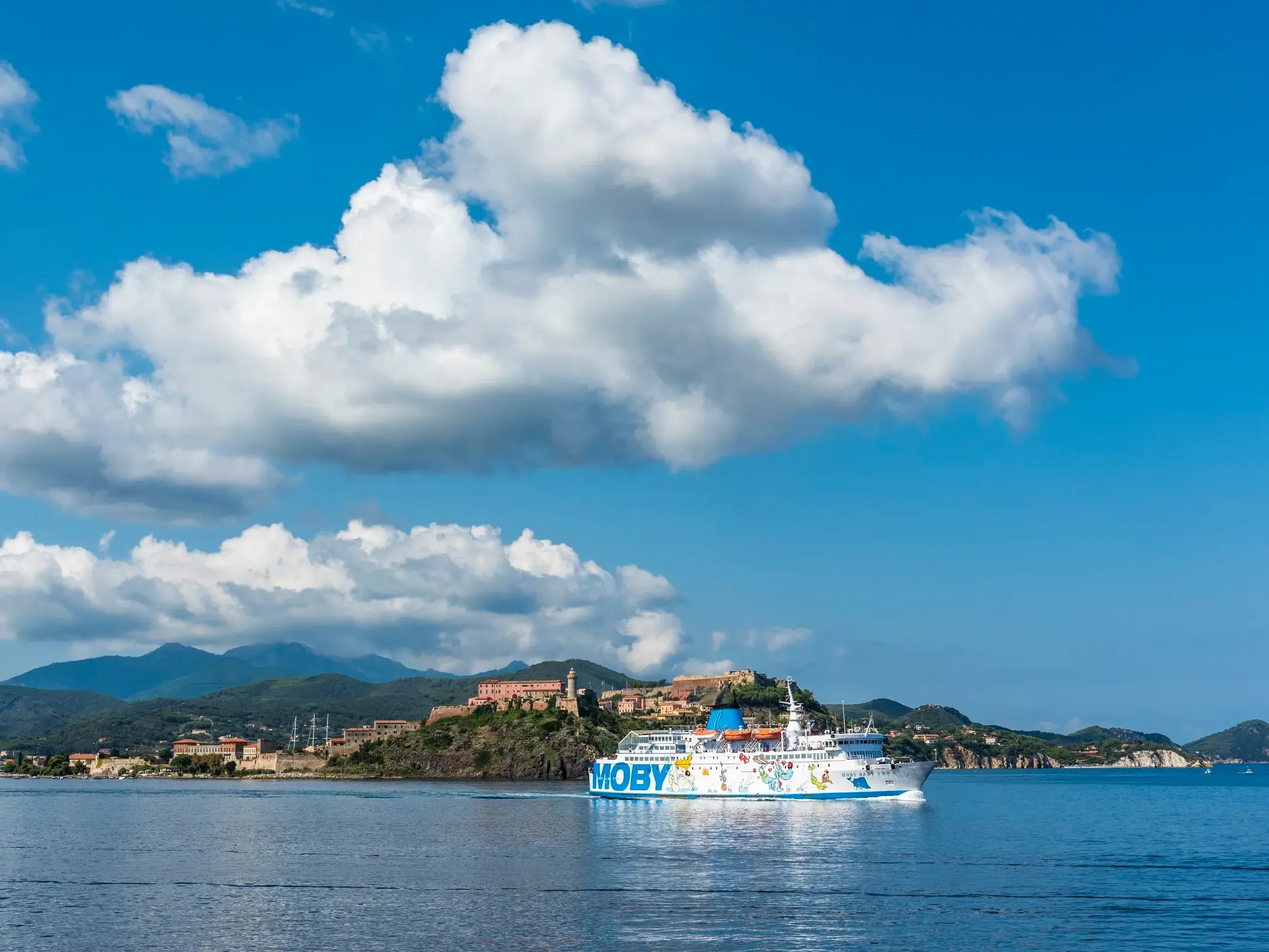 Eine Moby-Fähre fährt bei sonnigem Wetter an der Küste Elbas entlang, im Hintergrund die Hafenstadt Portoferraio.
