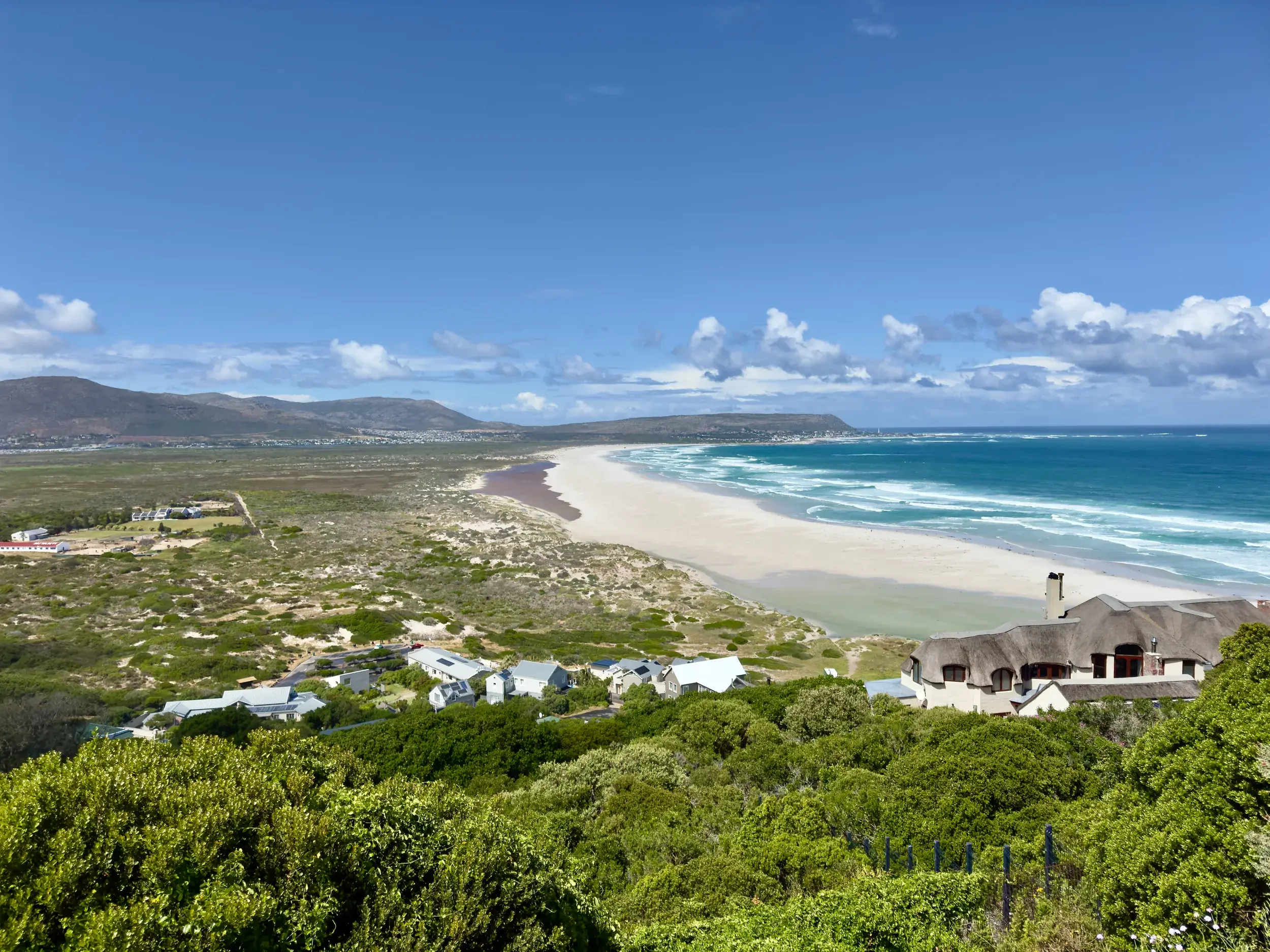 Panoramablick auf den langen, weißen Sandstrand von Noordhoek bei Kapstadt, umgeben von grünen Hügeln und dem blauen Ozean.