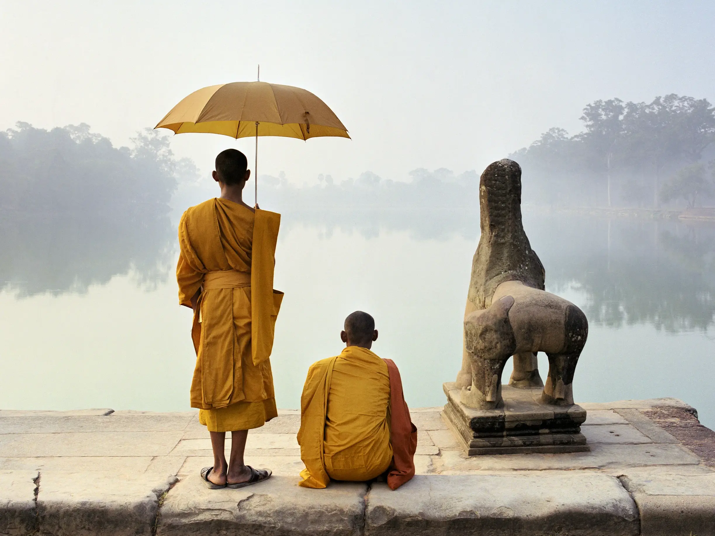 Zwei Mönche in safranfarbenen Roben an einem nebligen Seeufer in Angkor, Kambodscha, neben einer alten Löwenstatue.