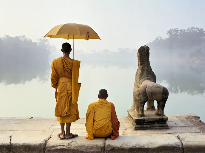 Zwei Mönche in safranfarbenen Roben an einem nebligen Seeufer in Angkor, Kambodscha, neben einer alten Löwenstatue.