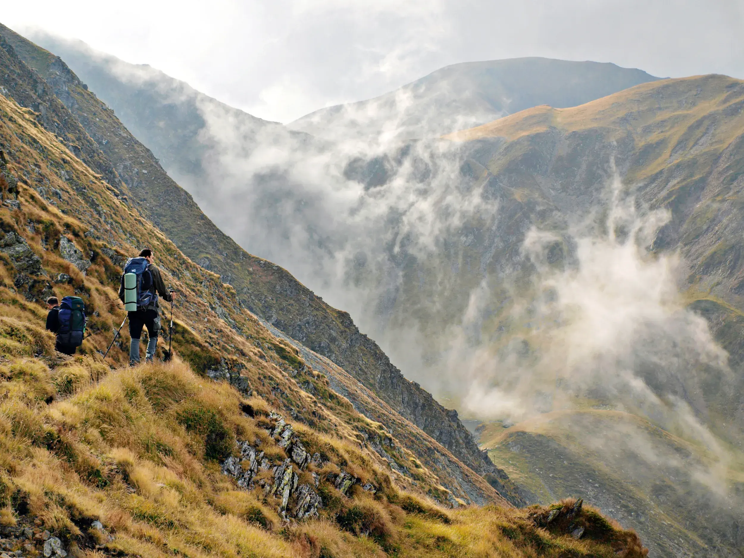 Zwei Trekker mit Rucksäcken auf einem schmalen Höhenweg durch das nebelverhangene Fagaras Gebirge in den rumänischen Karpaten.