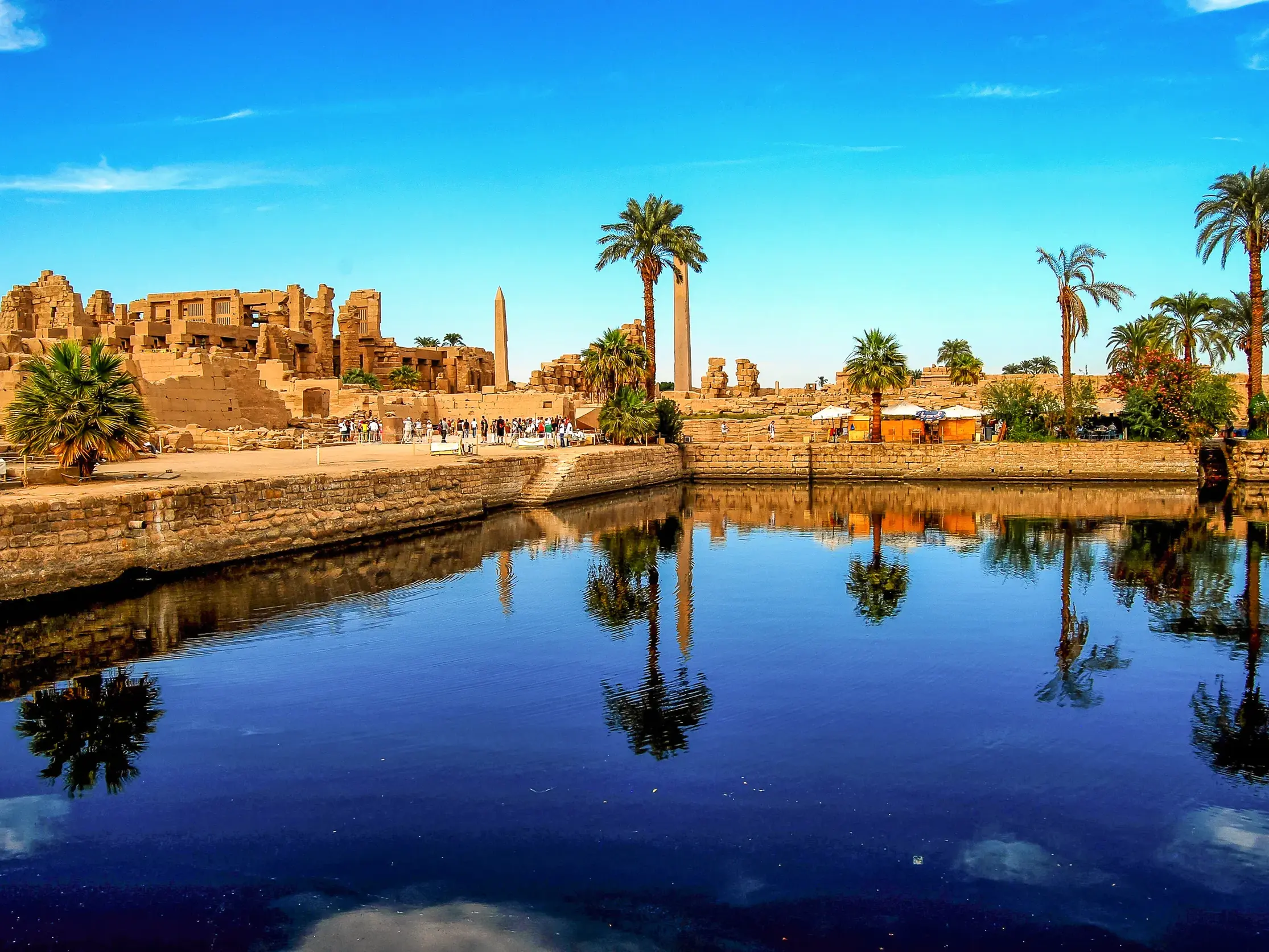 Der Heilige See in Karnak spiegelt die antiken Tempelruinen und Palmen unter blauem Himmel wider. Eine Reisegruppe erkundet die Anlage.