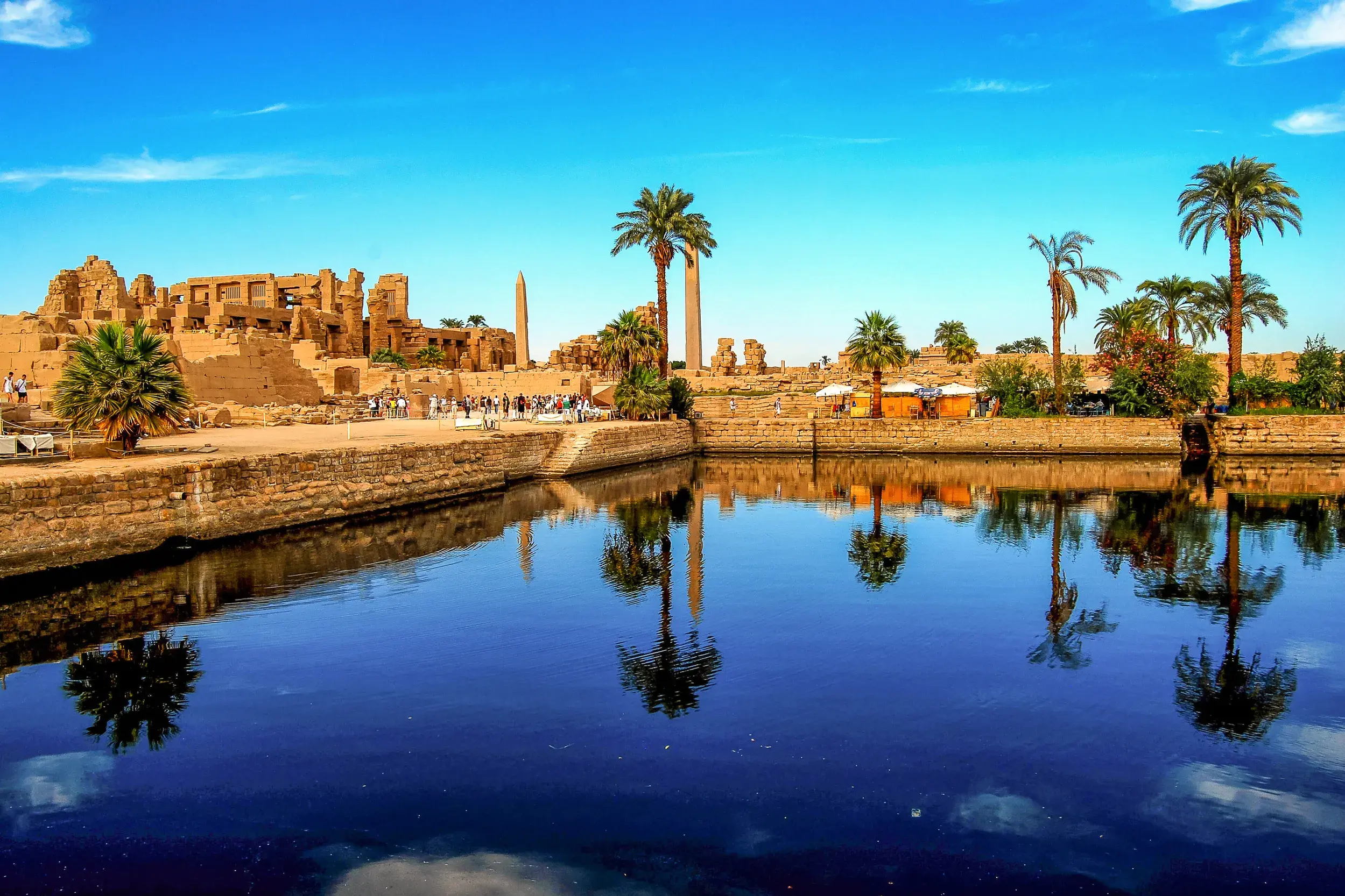 Der Heilige See in Karnak spiegelt die antiken Tempelruinen und Palmen unter blauem Himmel wider. Eine Reisegruppe erkundet die Anlage.