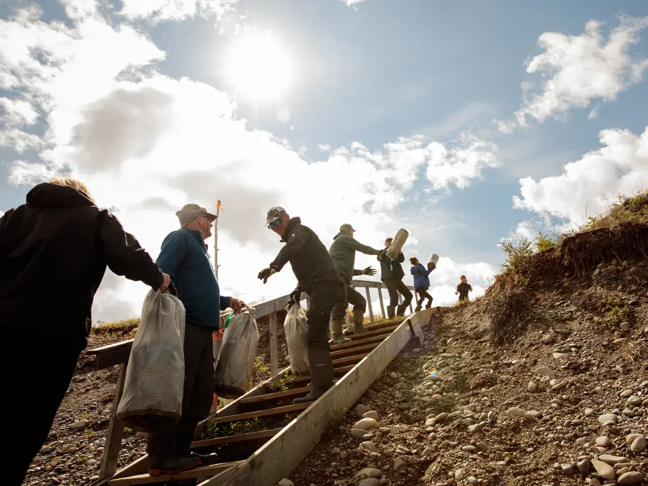 Hauser-Reisegruppe trägt bei Sonnenschein Packsäcke eine Holztreppe am steinigen Ufer des Yukon River in Kanada hinauf.