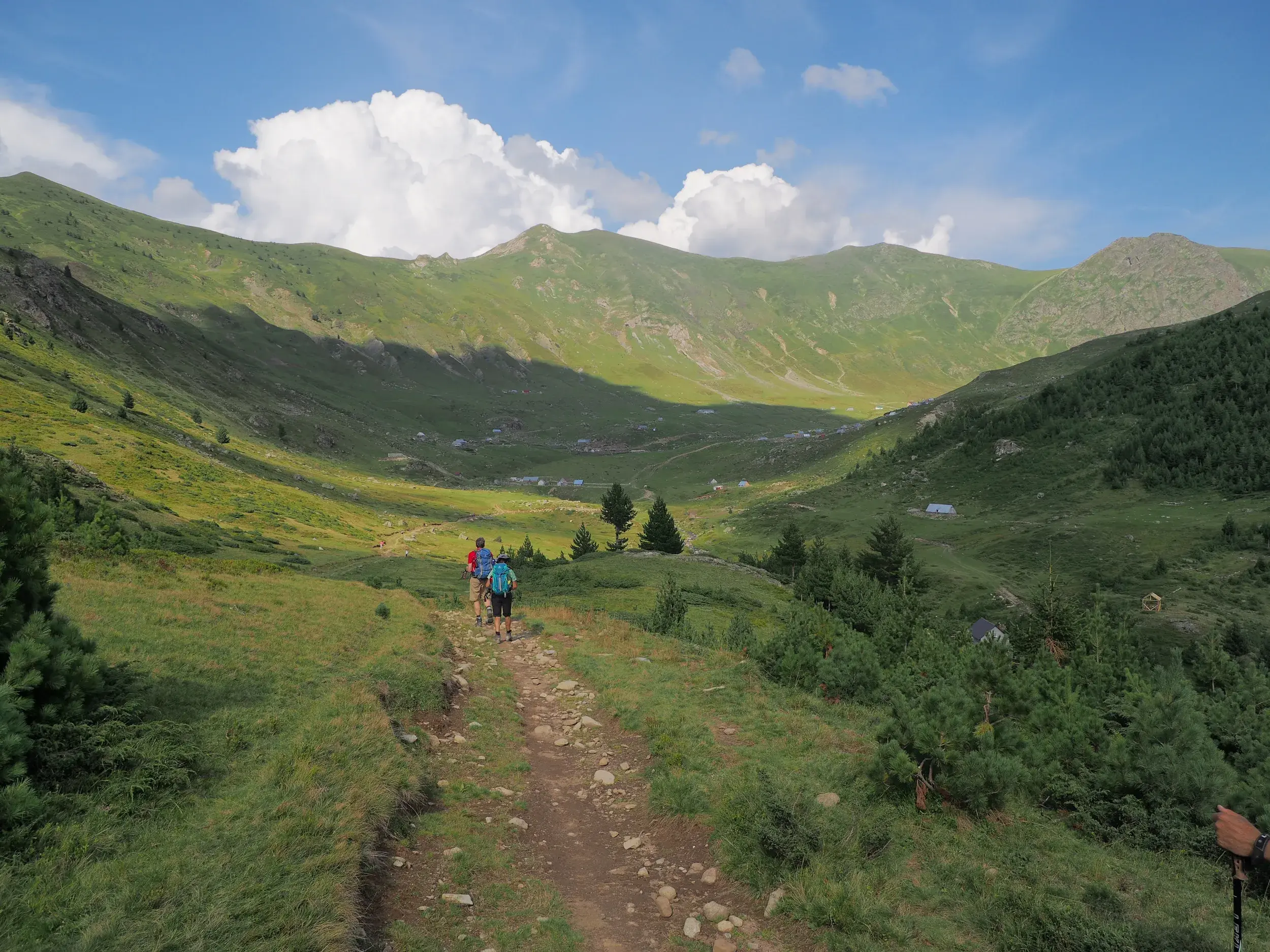 Wanderer mit Rucksäcken auf einem Pfad, der in ein weites, grünes Tal mit Berghütten auf dem Peaks of the Balkans-Trail führt.