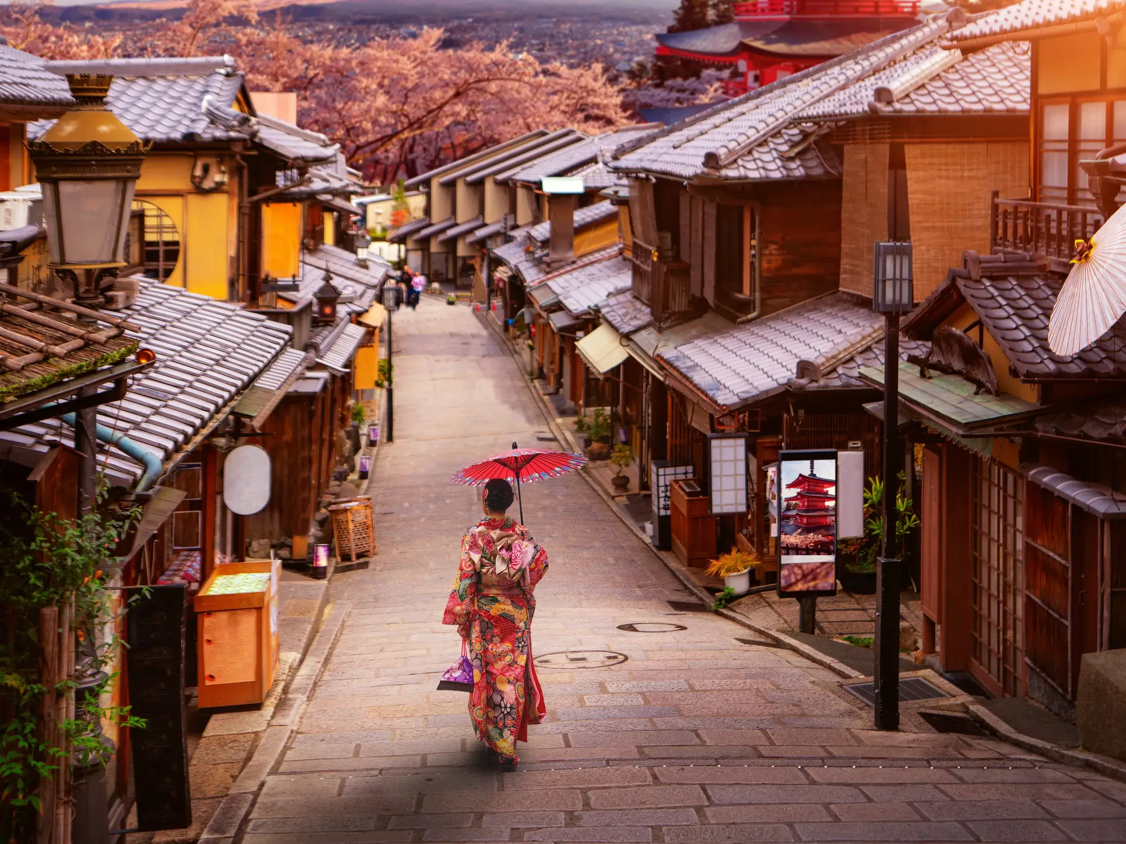 Frau im bunten Kimono mit rotem Schirm in einer gepflasterten Gasse in Kyoto, im Hintergrund Kirschblüten und eine Pagode.