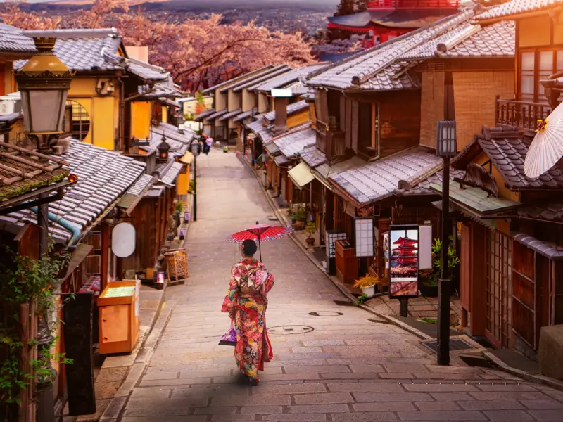 Frau im bunten Kimono mit rotem Schirm in einer gepflasterten Gasse in Kyoto, im Hintergrund Kirschblüten und eine Pagode.
