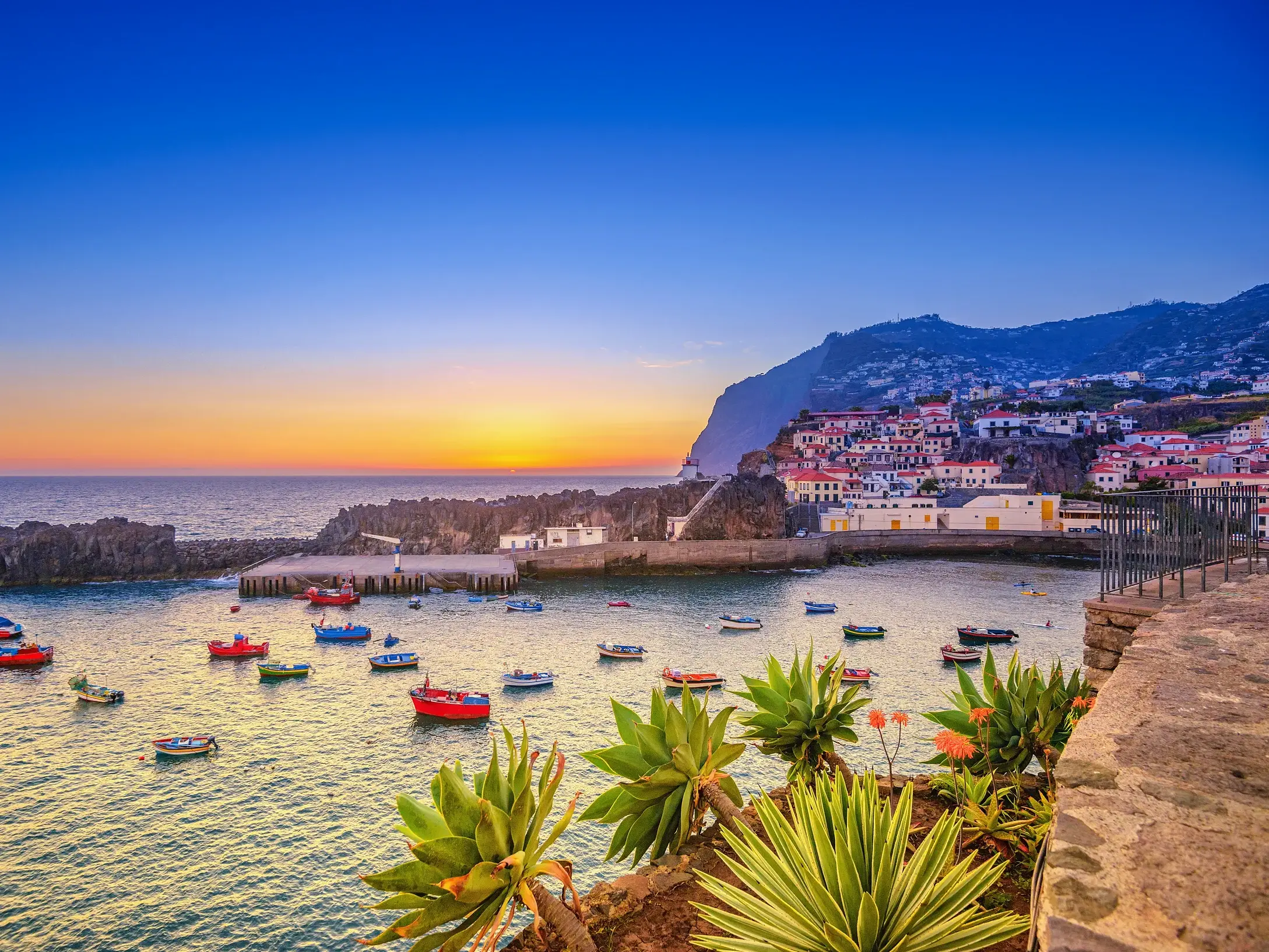 Malerische Bucht mit bunten Fischerbooten vor einem Küstendorf auf Madeira bei Sonnenuntergang über dem Atlantik.