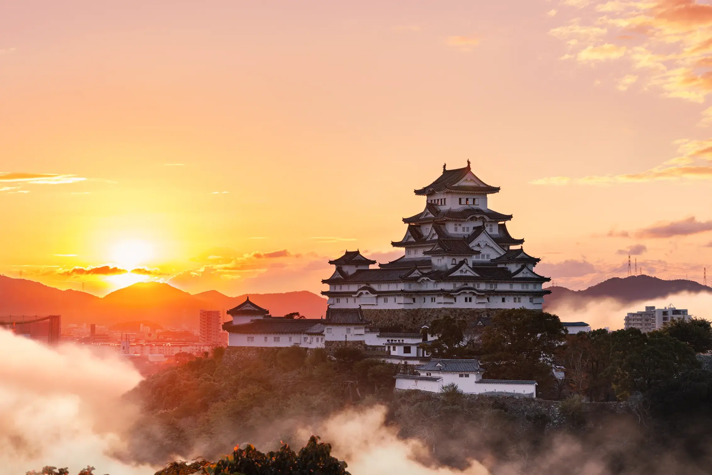 Die strahlend weiße Burg Himeji in Japan thront bei Sonnenaufgang über einem Meer aus Wolken und Bergen am Horizont.