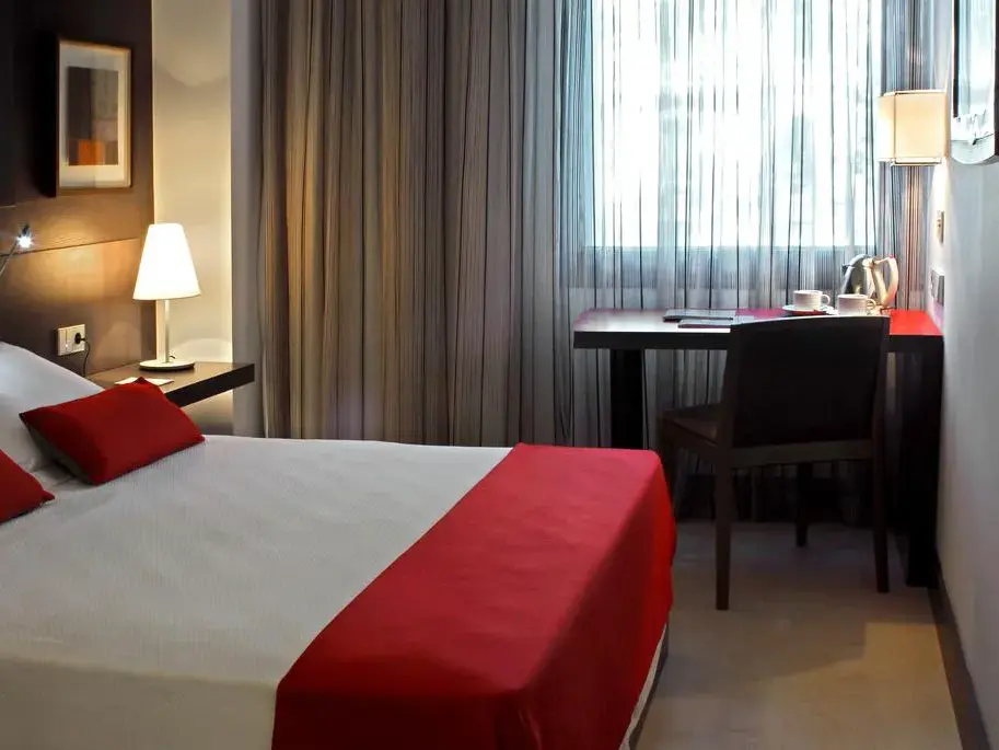Modern eingerichtetes Doppelzimmer im Grupotel Gran Vía 678 mit gemachtem Bett, roten Akzenten und Schreibtisch am Fenster.