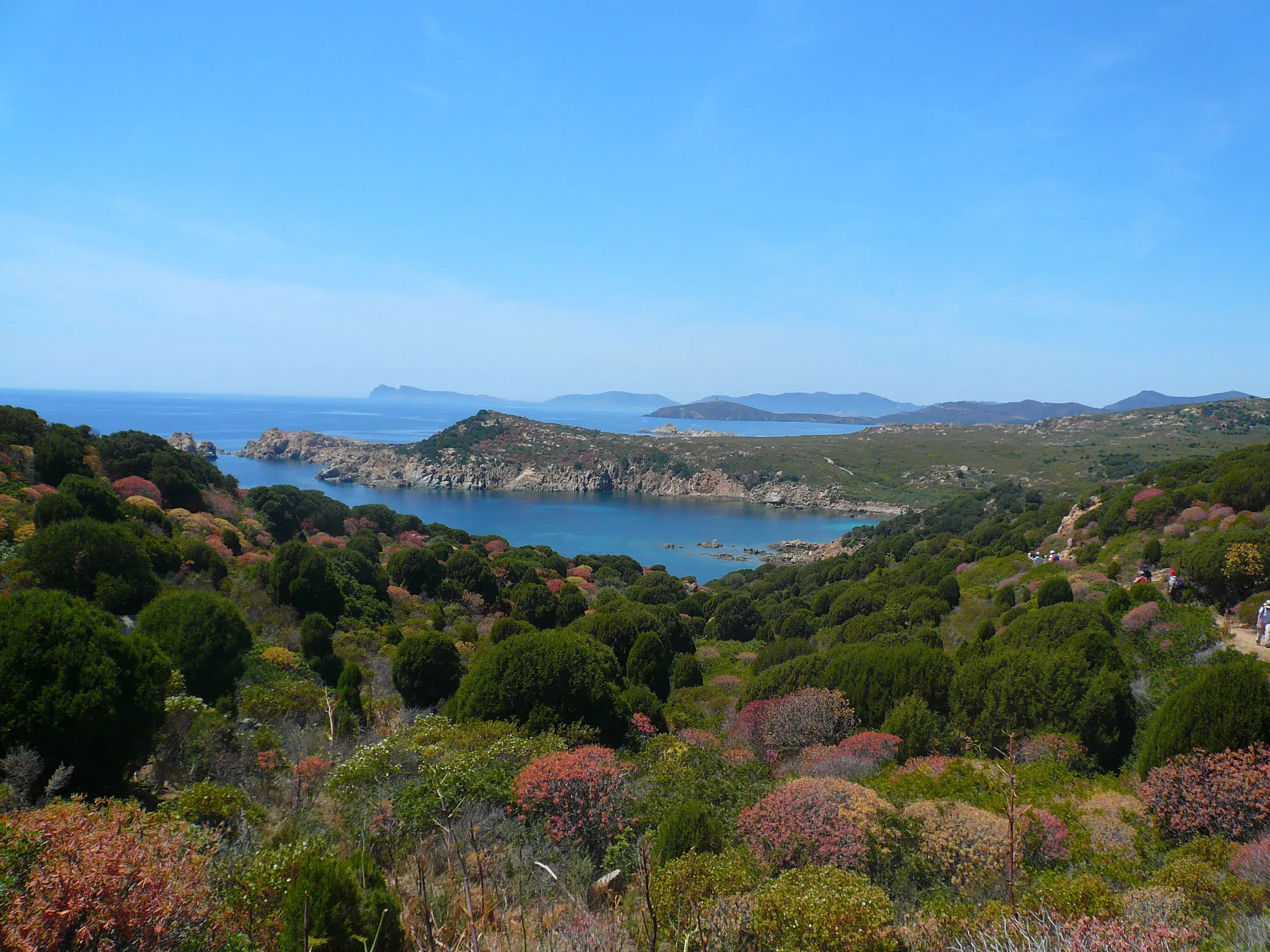 Wandergruppe auf einem Küstenpfad am Capo Testa auf Sardinien mit Panoramablick über die Macchia auf eine türkisblaue Bucht.
