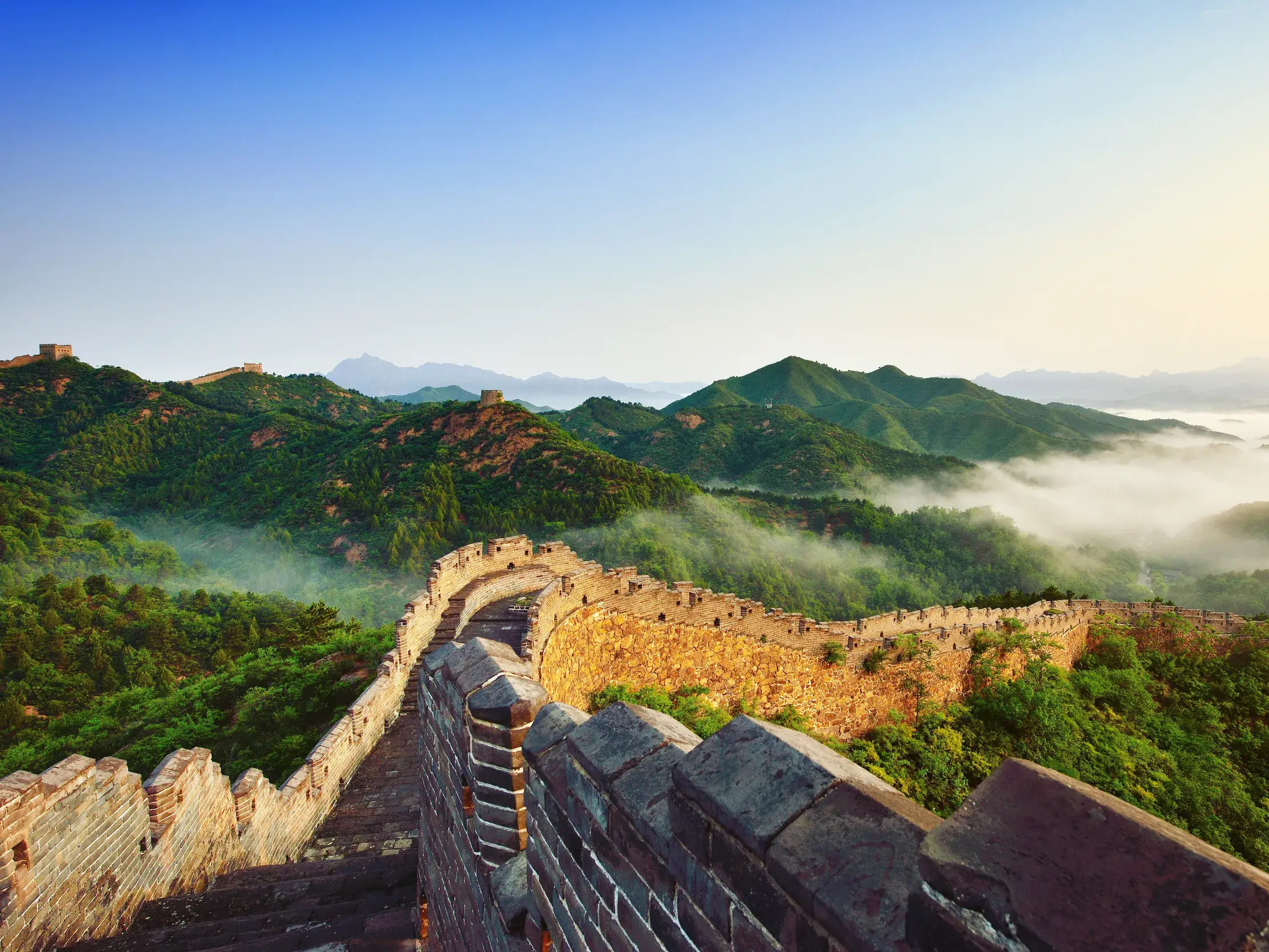 Das UNESCO-Welterbe Große Mauer schlängelt sich bei Sonnenaufgang über grüne, nebelverhangene Berge in China.