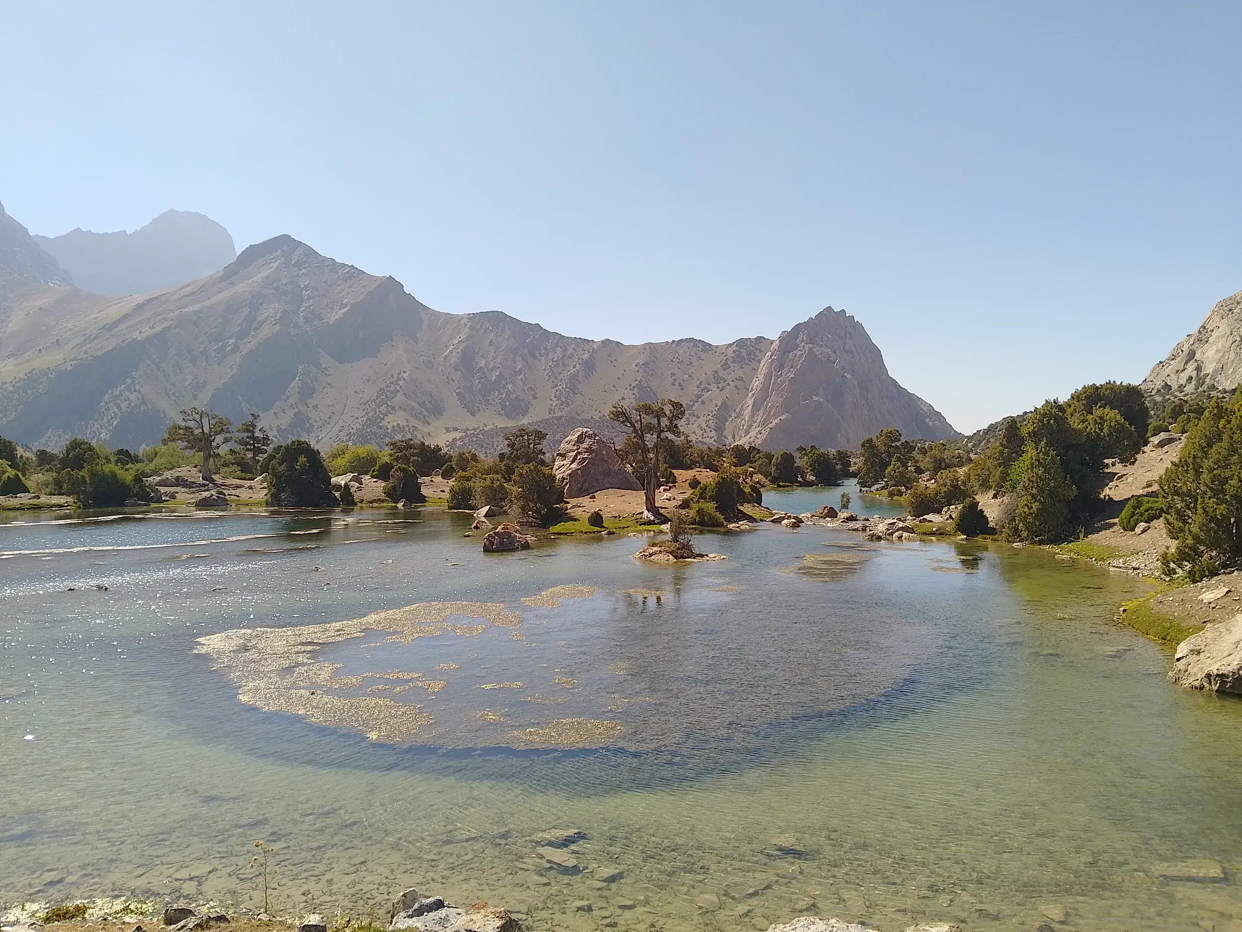 Glasklarer Bergsee im Fan-Gebirge in Tadschikistan, umgeben von grünen Bäumen und den hohen, schroffen Berggipfeln.