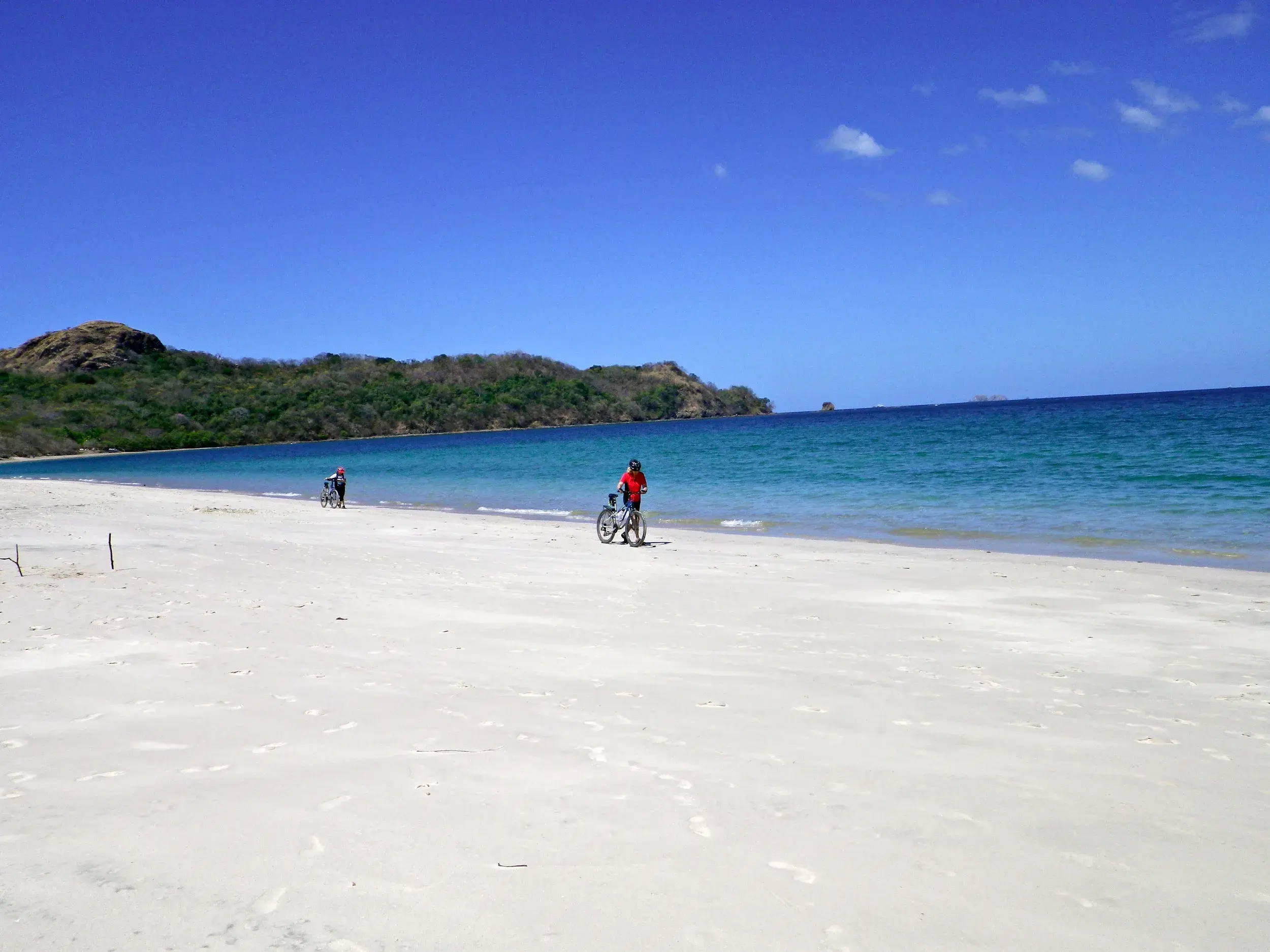 Zwei Personen erkunden mit Fahrrädern einen einsamen, weißen Sandstrand am Pazifik in Costa Rica bei strahlendem Sonnenschein.