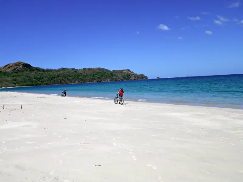 Zwei Personen erkunden mit Fahrrädern einen einsamen, weißen Sandstrand am Pazifik in Costa Rica bei strahlendem Sonnenschein.