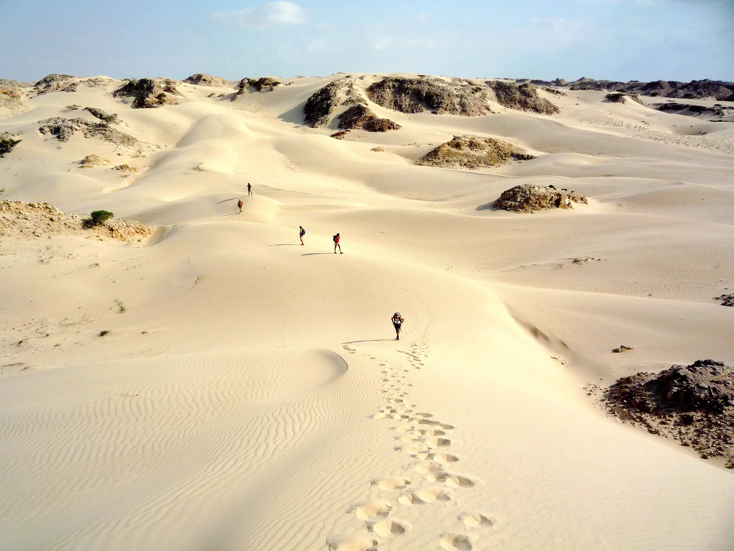 Eine kleine Wandergruppe beim Trekking durch die endlosen, weißen Sanddünen der Wüste Al-Khaluf im Oman bei Sonnenschein.