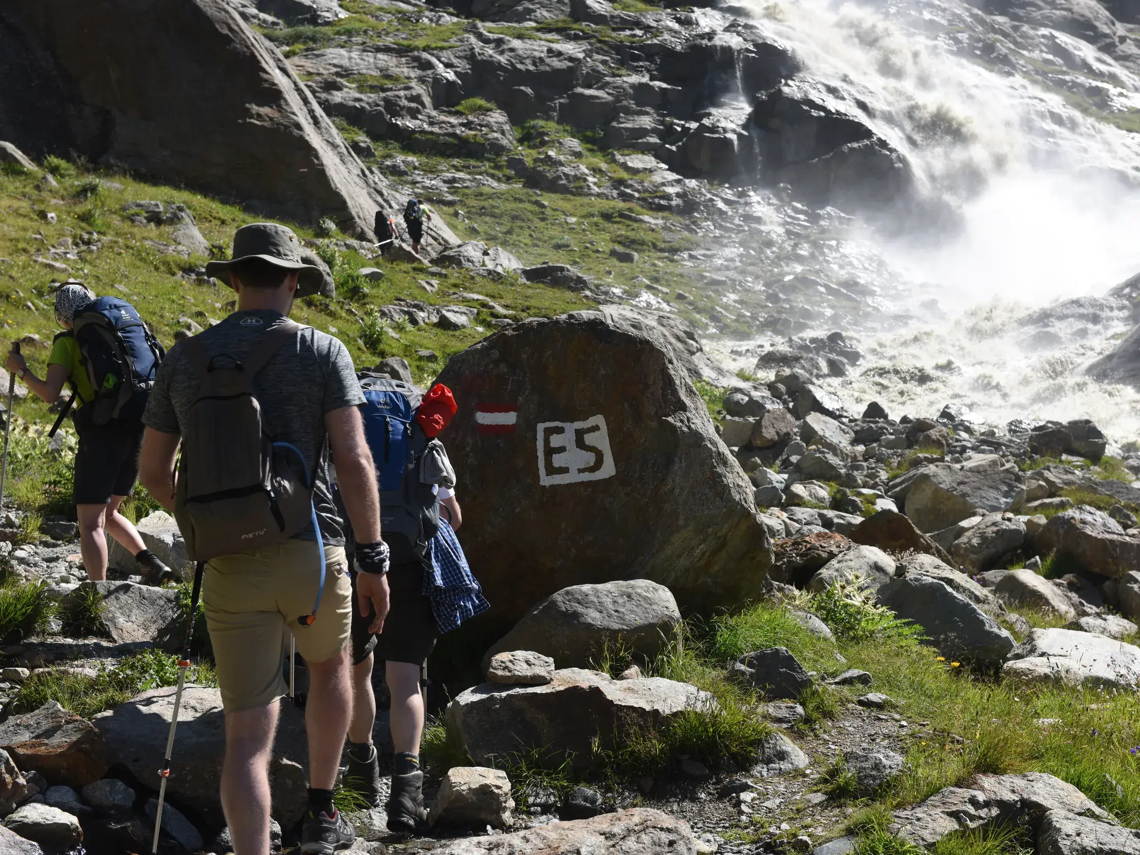 Eine Wandergruppe folgt der E5-Wegmarkierung auf einem steinigen Pfad entlang eines tosenden Wasserfalls in den Alpen.