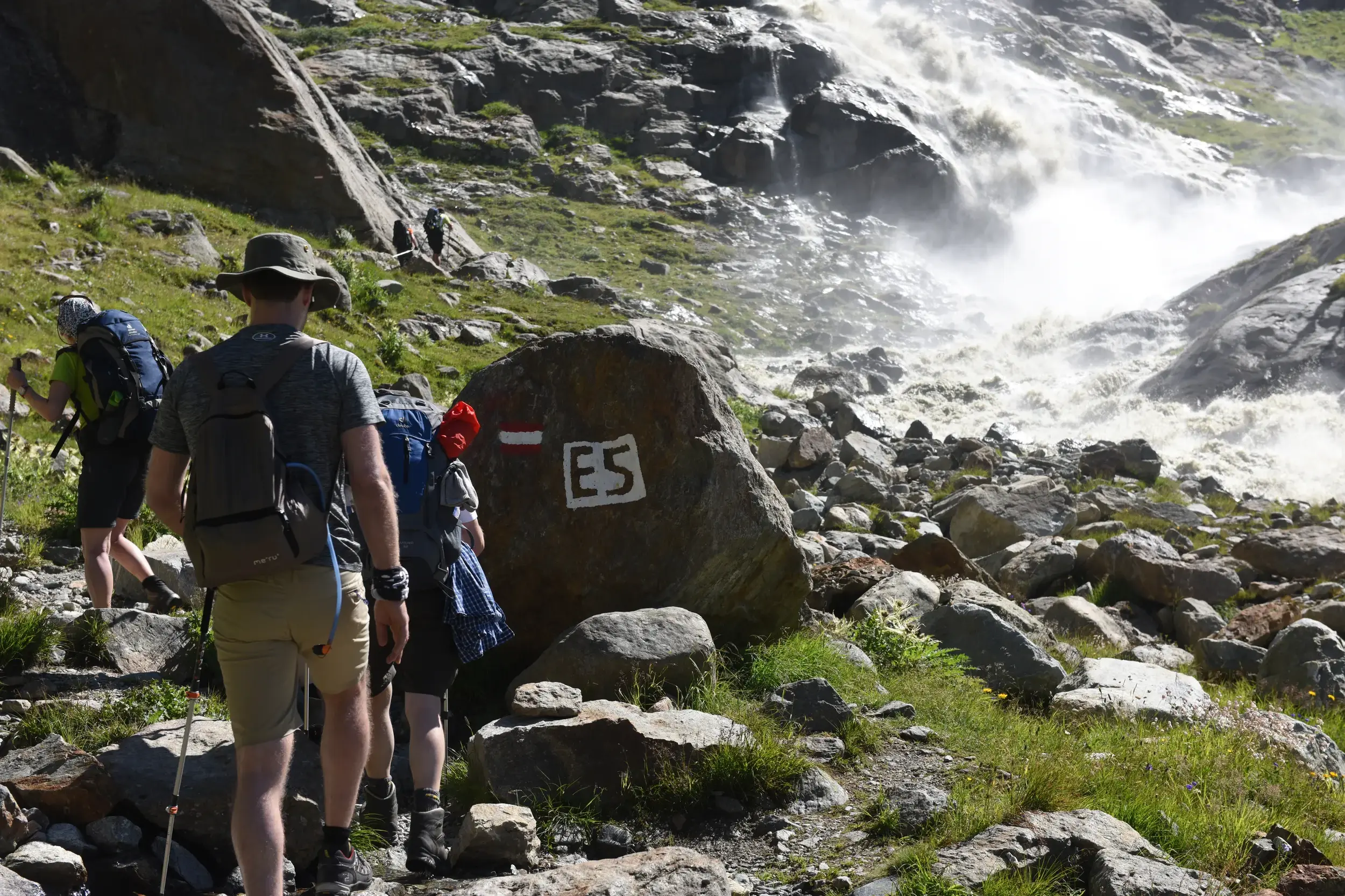Eine Wandergruppe folgt der E5-Wegmarkierung auf einem steinigen Pfad entlang eines tosenden Wasserfalls in den Alpen.