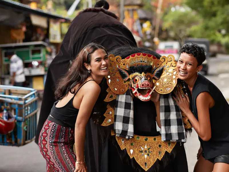 Zwei junge Frauen erleben auf einer Rundreise in Bali eine kulturelle Begegnung und posieren lachend mit einer Barong-Maske.