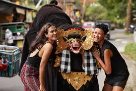 Zwei junge Frauen erleben auf einer Rundreise in Bali eine kulturelle Begegnung und posieren lachend mit einer Barong-Maske.