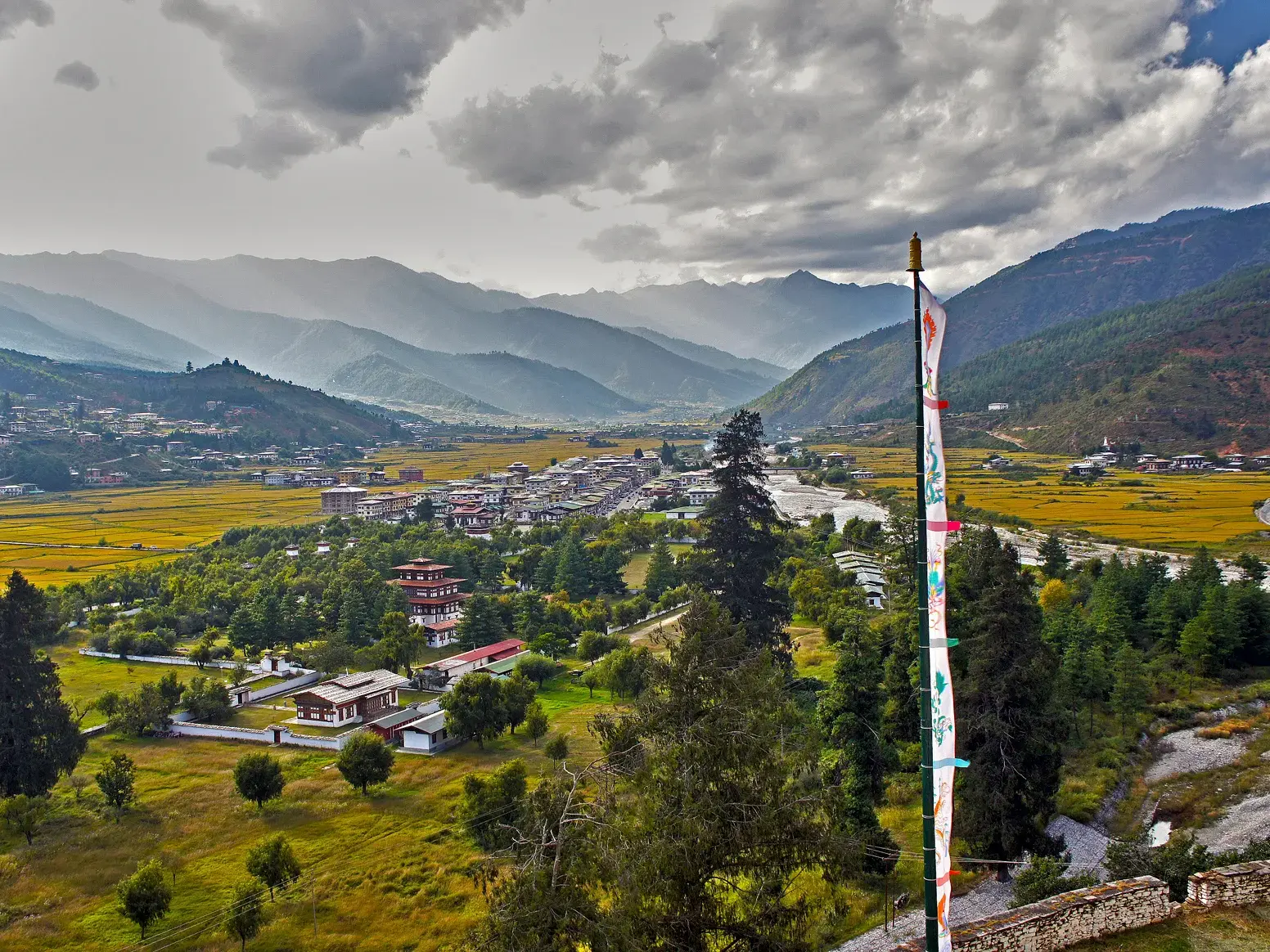 Panoramablick über das Paro-Tal in Bhutan mit Gebetsfahne, goldenen Reisfeldern und der Himalaya-Bergkette im Hintergrund.