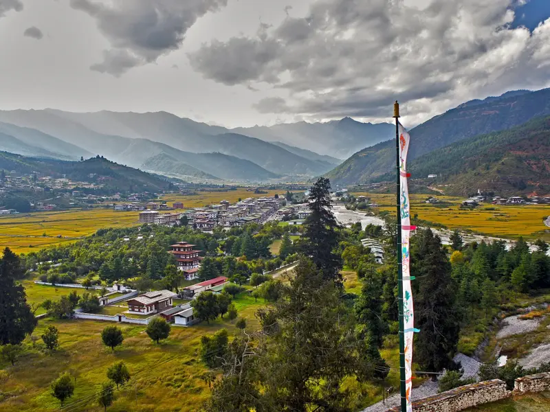 Panoramablick über das Paro-Tal in Bhutan mit Gebetsfahne, goldenen Reisfeldern und der Himalaya-Bergkette im Hintergrund.