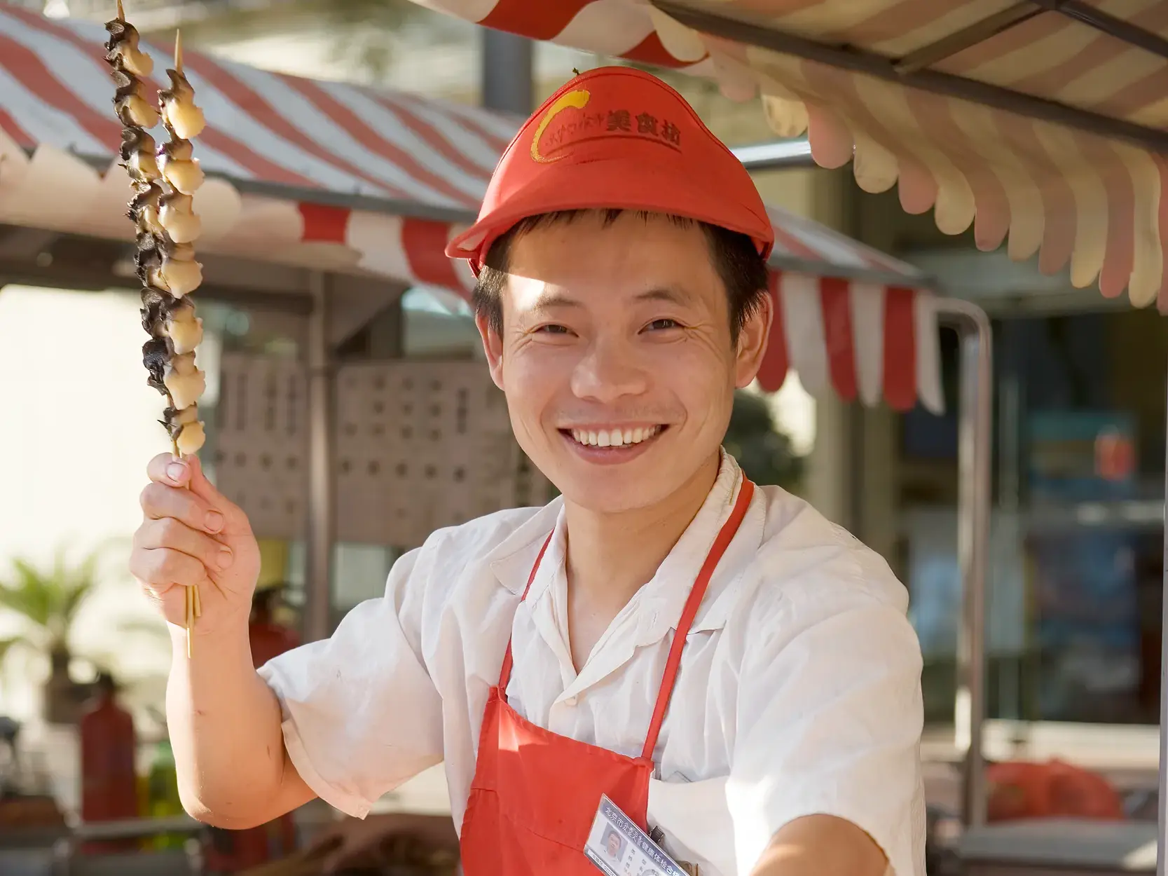Ein freundlicher chinesischer Verkäufer in roter Schürze und Kappe präsentiert an seinem Stand lächelnd einen Spieß mit gegrillten Meeresschnecken.