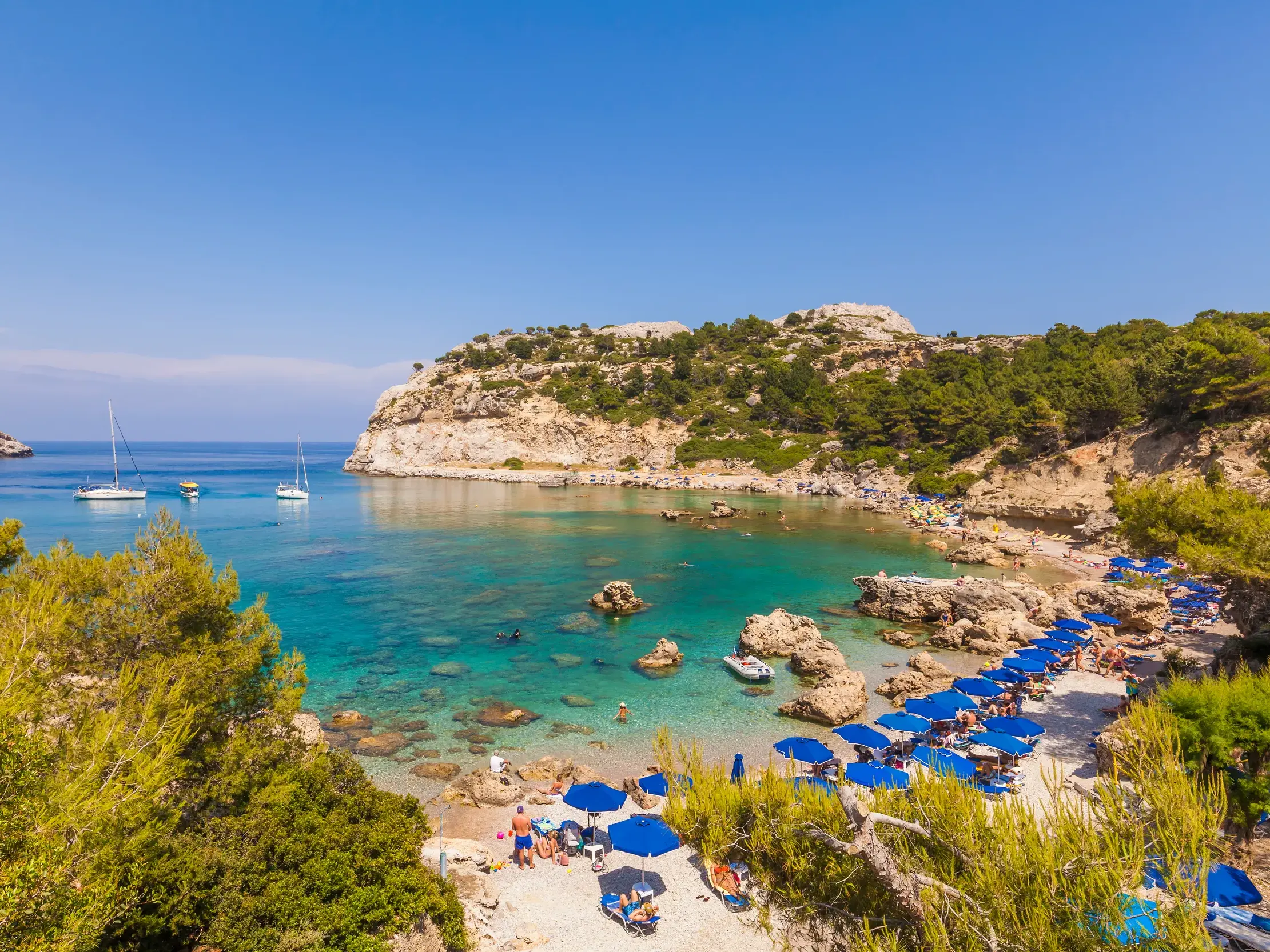Sonnige Felsenbucht auf Rhodos mit türkisem Ägäis-Wasser, Segelbooten und Badestrand mit blauen Sonnenschirmen.
