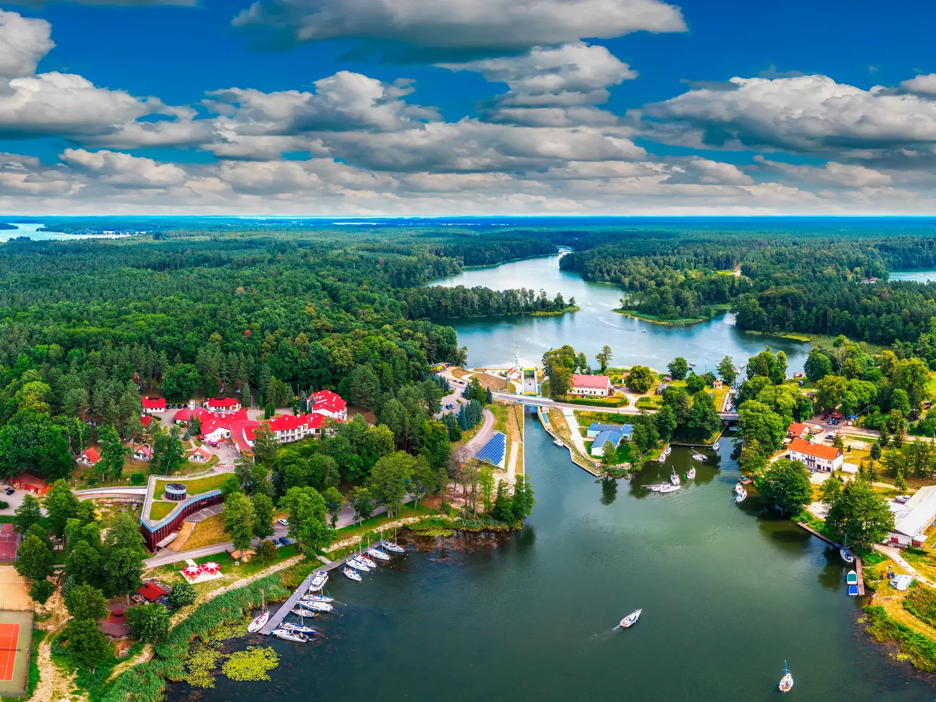 Luftaufnahme der Masurischen Seenplatte in Polen mit dem Ort Mikolajki, vielen Seen, Wäldern und Booten unter blauem Himmel.