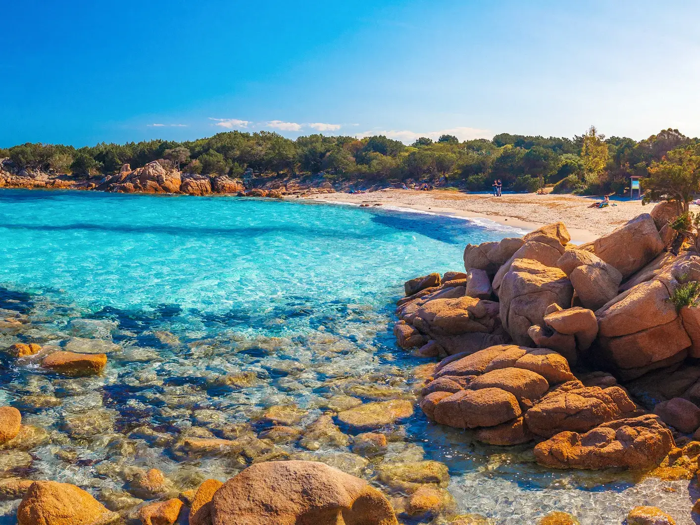 Kristallklares, türkises Wasser an der felsigen Küste Sardiniens umspült einen Sandstrand unter strahlend blauem Himmel.