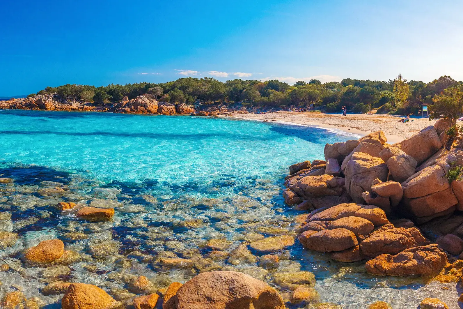 Kristallklares, türkises Wasser an der felsigen Küste Sardiniens umspült einen Sandstrand unter strahlend blauem Himmel.