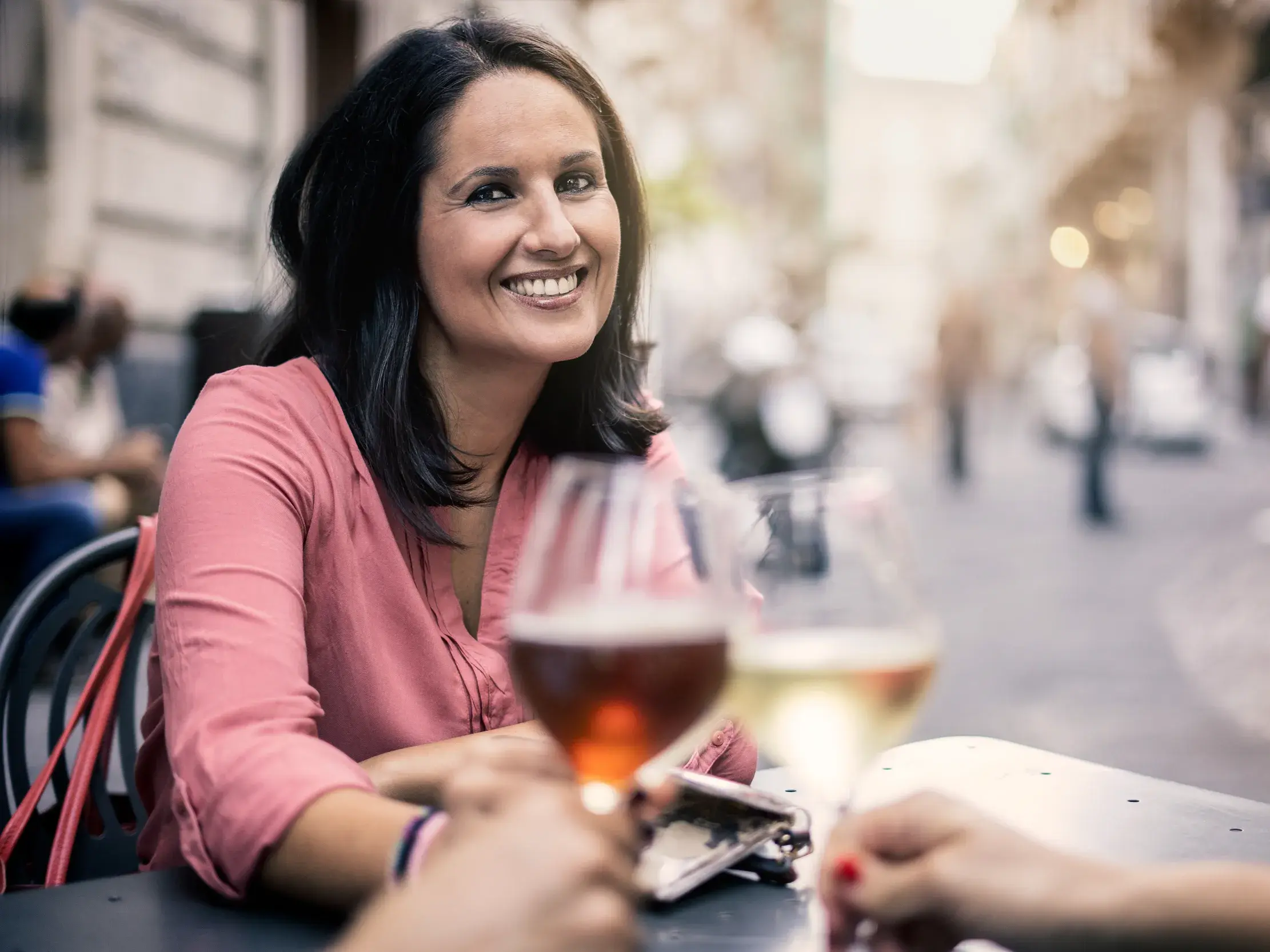 Eine lächelnde Frau in einem rosa Oberteil genießt ein Glas Wein in einem belebten Straßencafé in der Altstadt von Bilbao.