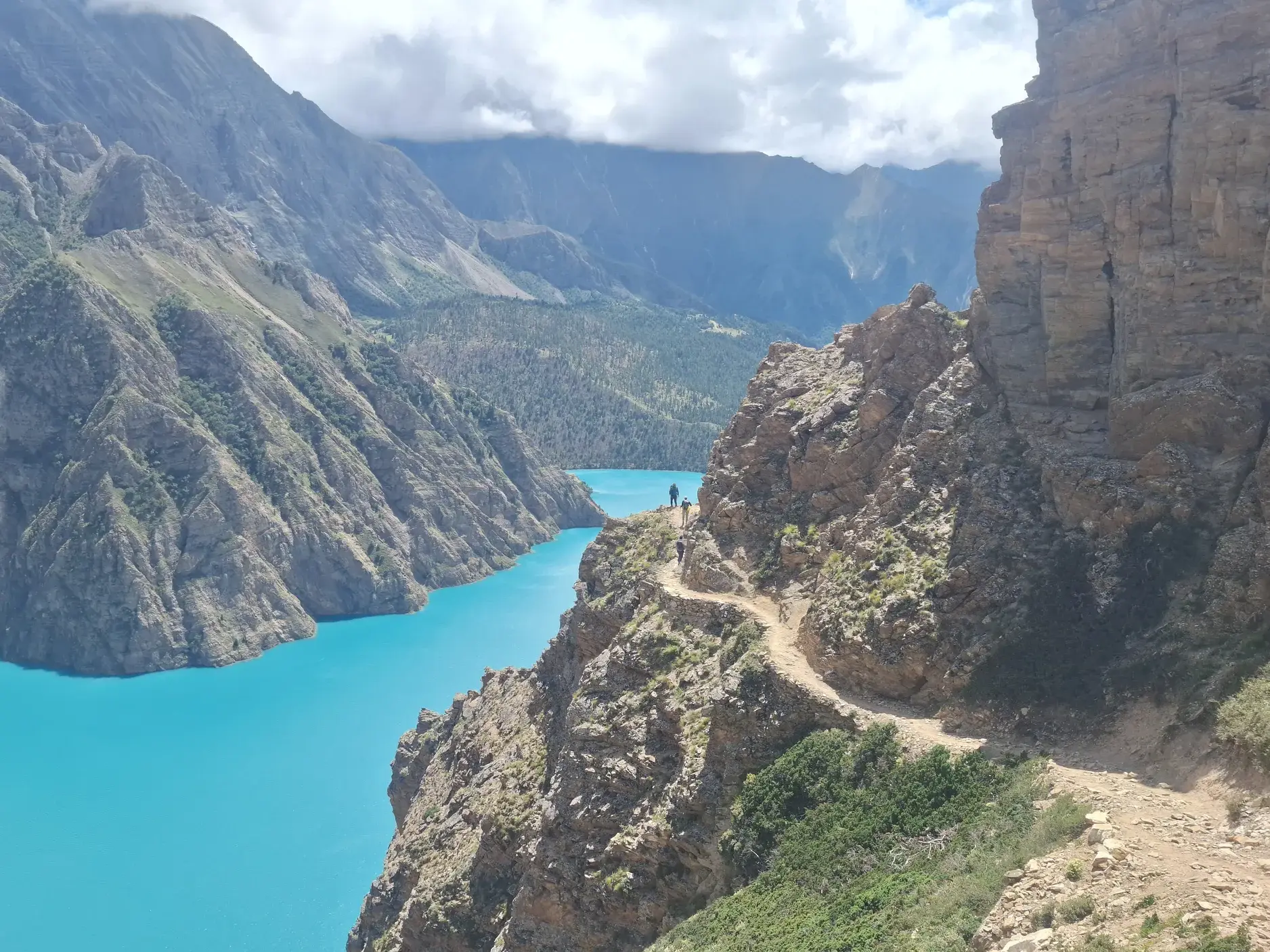 Zwei Wanderer auf einem schmalen Felsenpfad hoch über dem türkisfarbenen Phoksundo-See in der abgelegenen Dolpo-Region, Nepal.