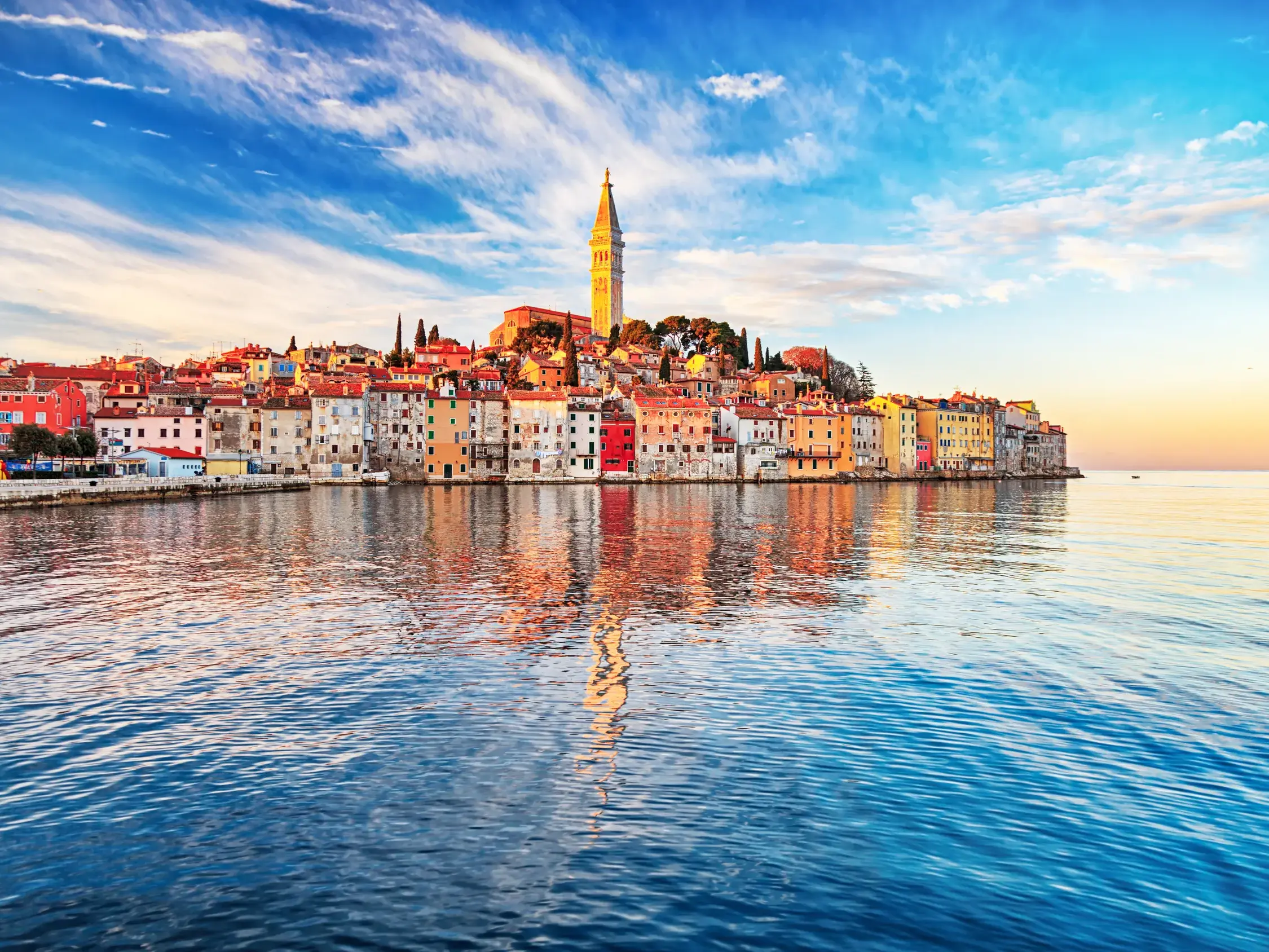 Die Küstenstadt Piran in Slowenien bei Sonnenuntergang. Die bunten Häuser der Altstadt spiegeln sich im ruhigen Wasser der Adria.