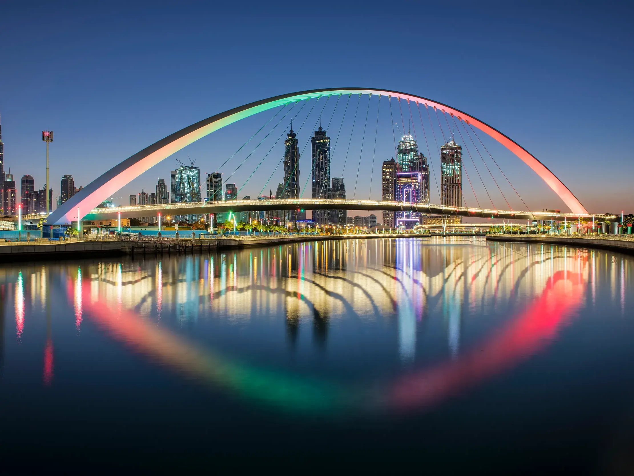 Die bunt beleuchtete Dubai Water Canal Bridge vor der nächtlichen Skyline mit dem Burj Khalifa, deren Lichter sich im Wasser spiegeln.